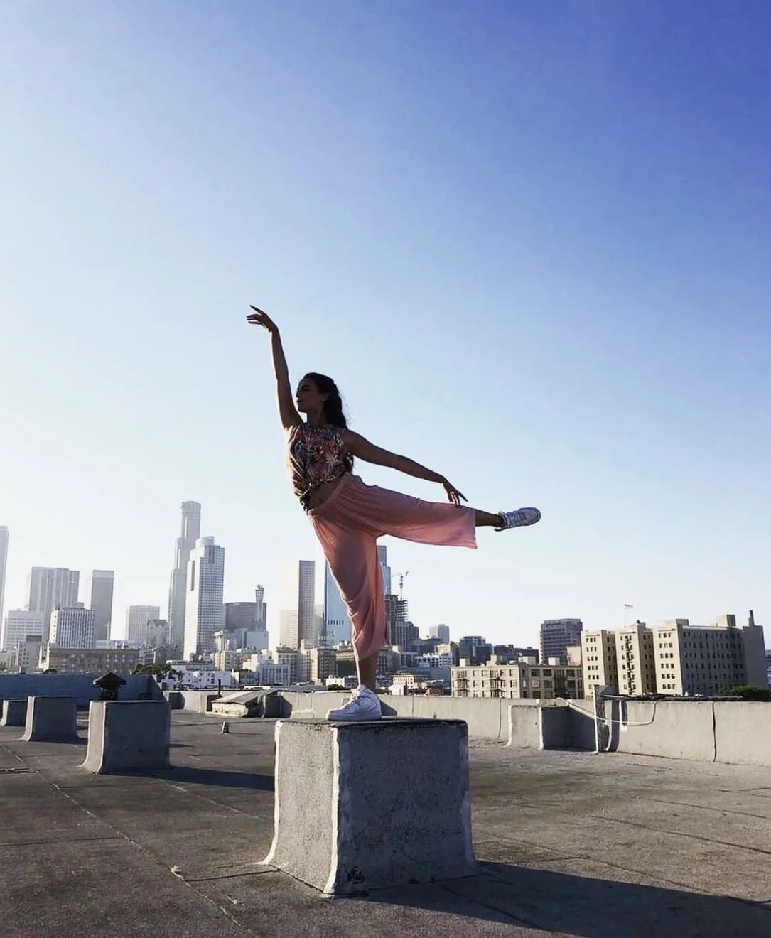A woman in a pink and floral outfit doing ballet pose on a rooftop with a city skyline in the background at sunset.
