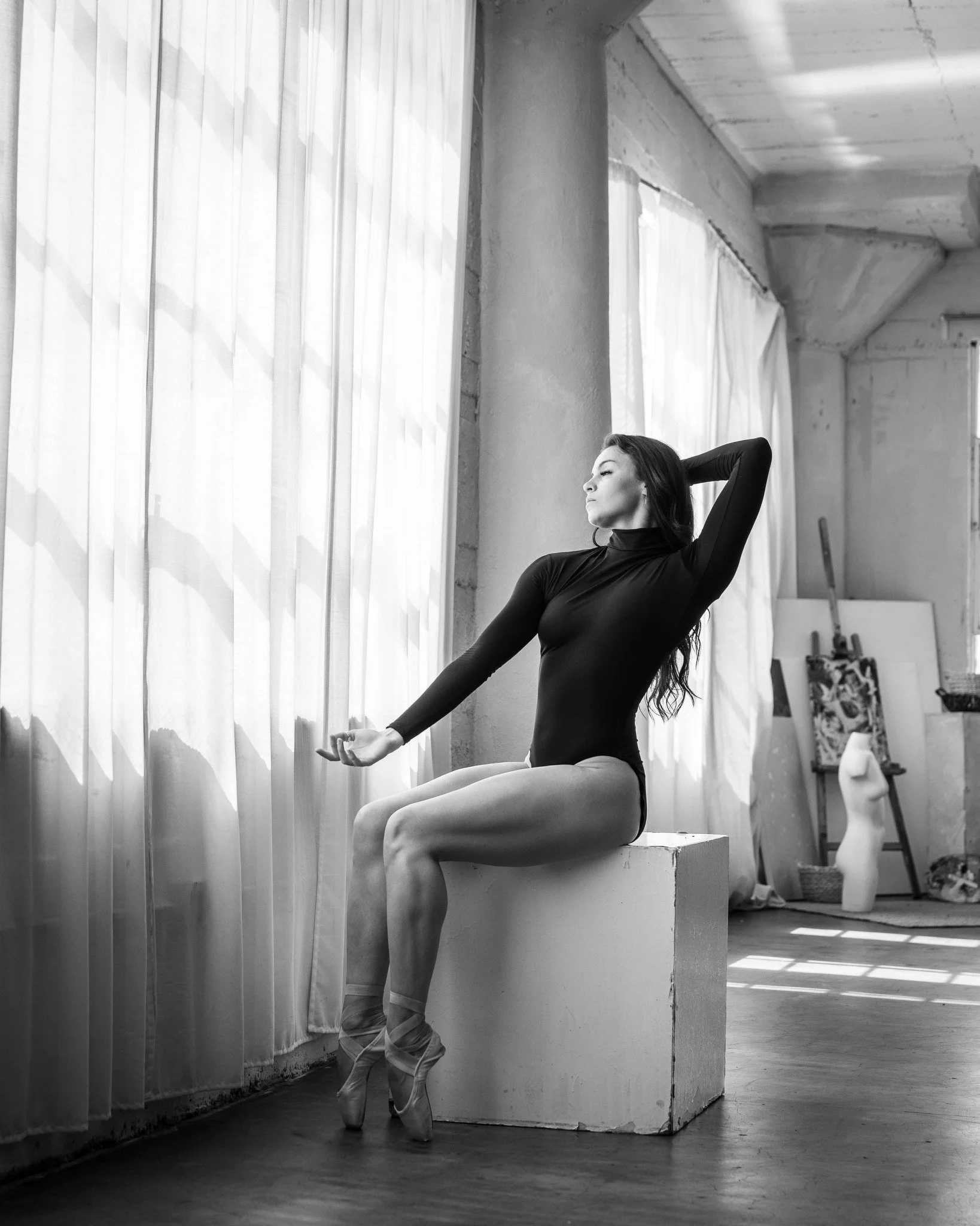 Ballet dancer in black leotard sitting on a block, stretching near a large window with sheer curtains in a dance studio.