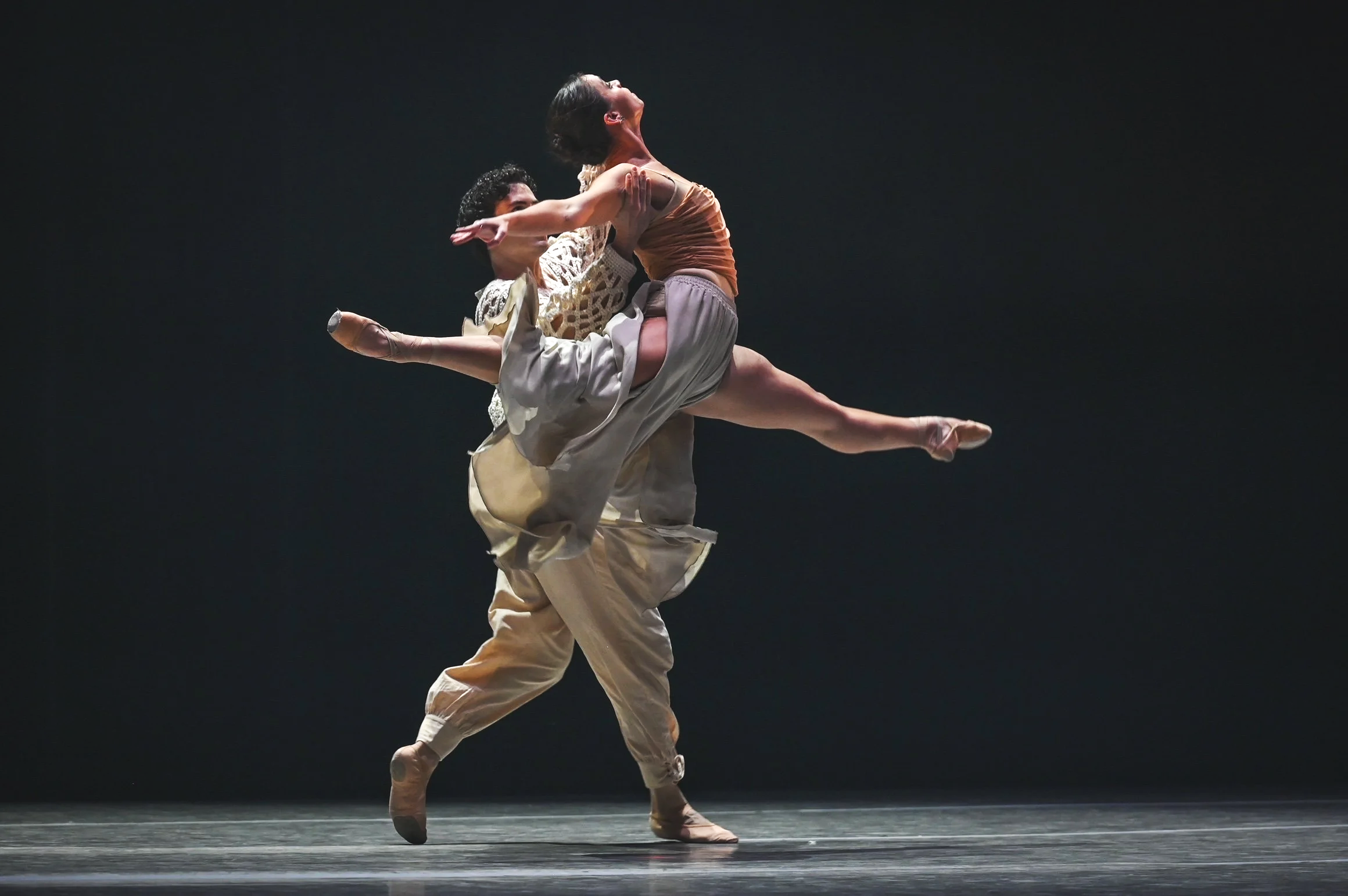 Two ballet dancers performing on stage with a black background, one lifting the other in an elegant pose.