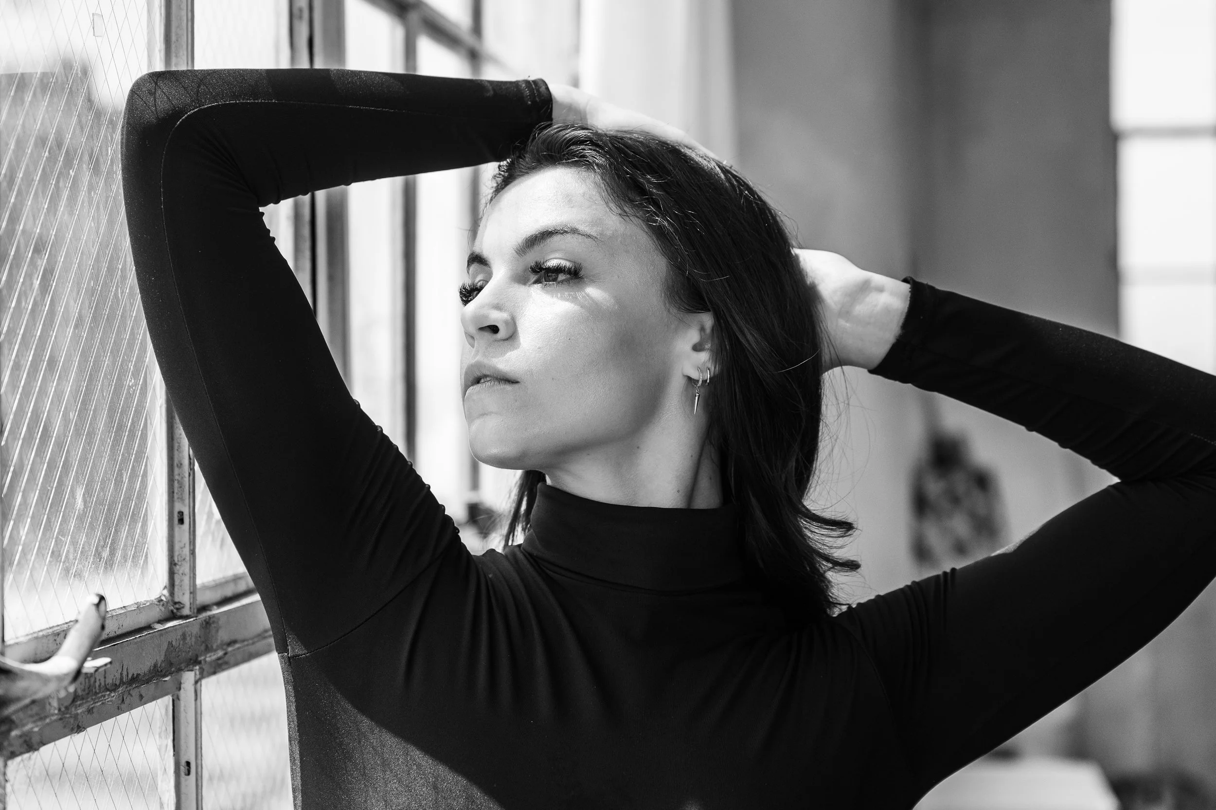 Black and white photo of a woman with dark hair posing with her arms behind her head, gazing thoughtfully to the side, wearing a long sleeve black top, near a window with industrial-style metal framing.