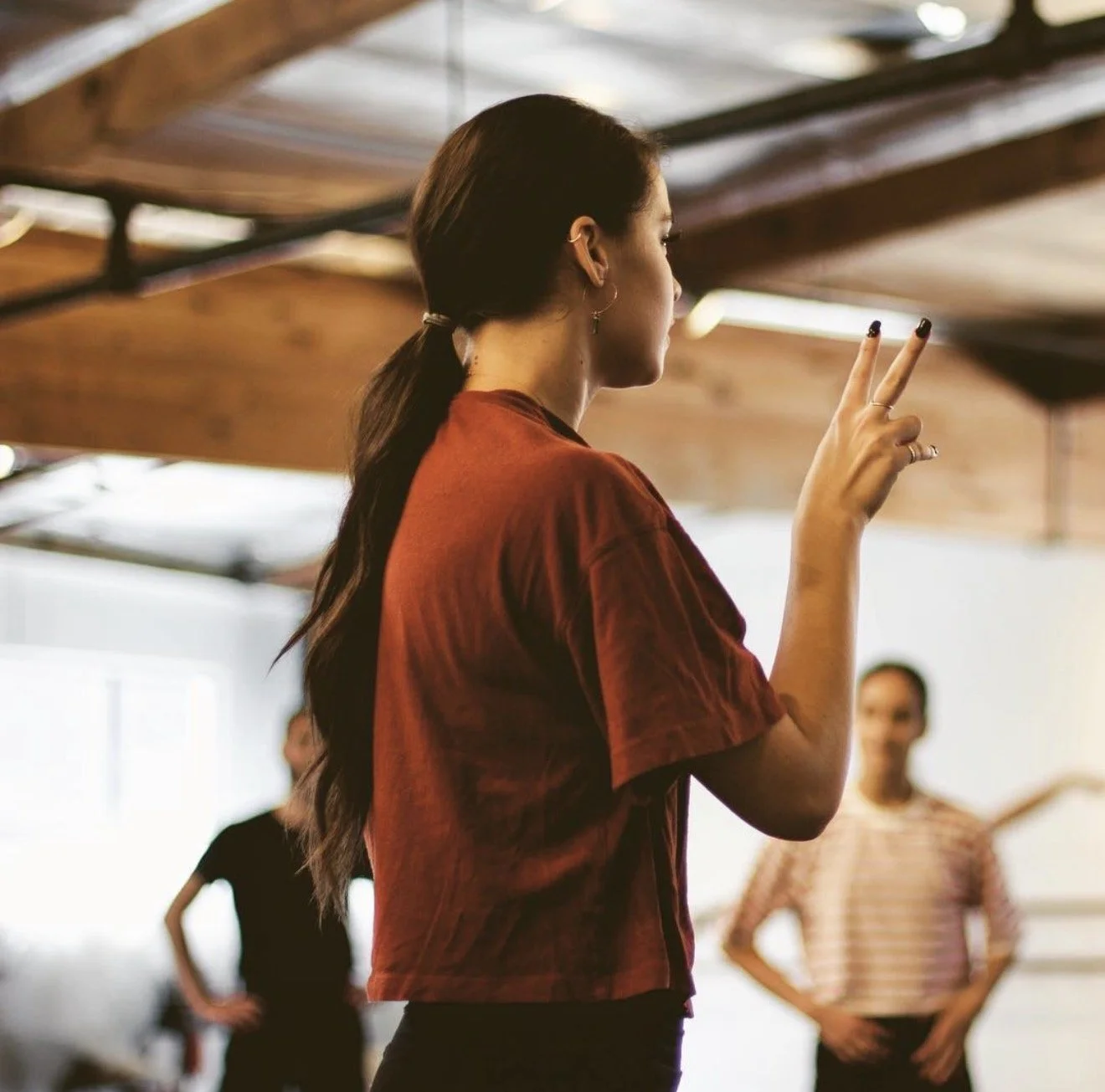 A young woman with long dark hair tied in a ponytail, wearing a red top, making a peace sign with her right hand in a dance or exercise studio.