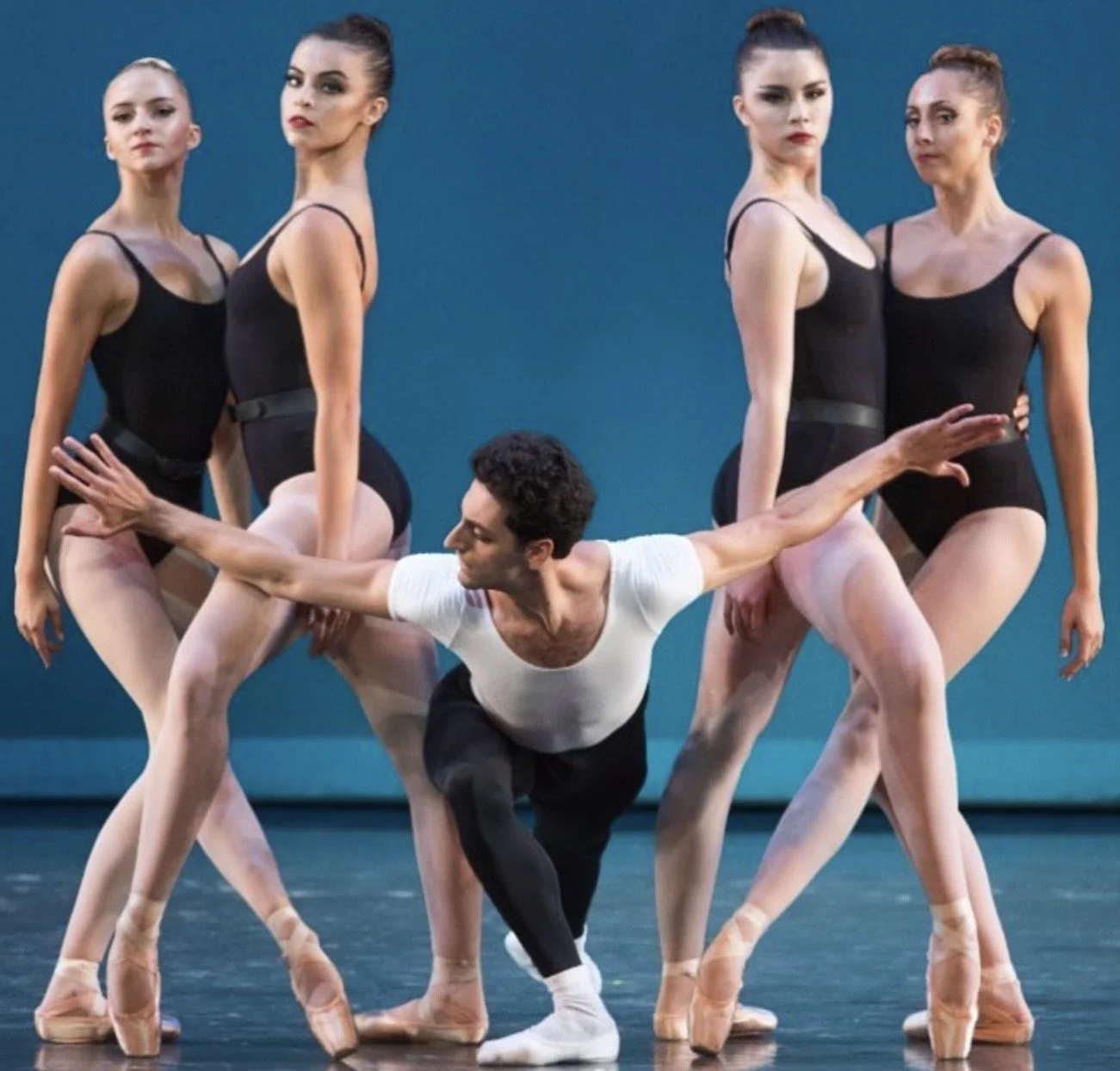 A ballet dancer with short dark hair is performing on stage with four women in black leotards and ballet slippers standing around him. The dancer is in a deep plié with one arm extended and smiling, while the women are posing in ballet positions agai