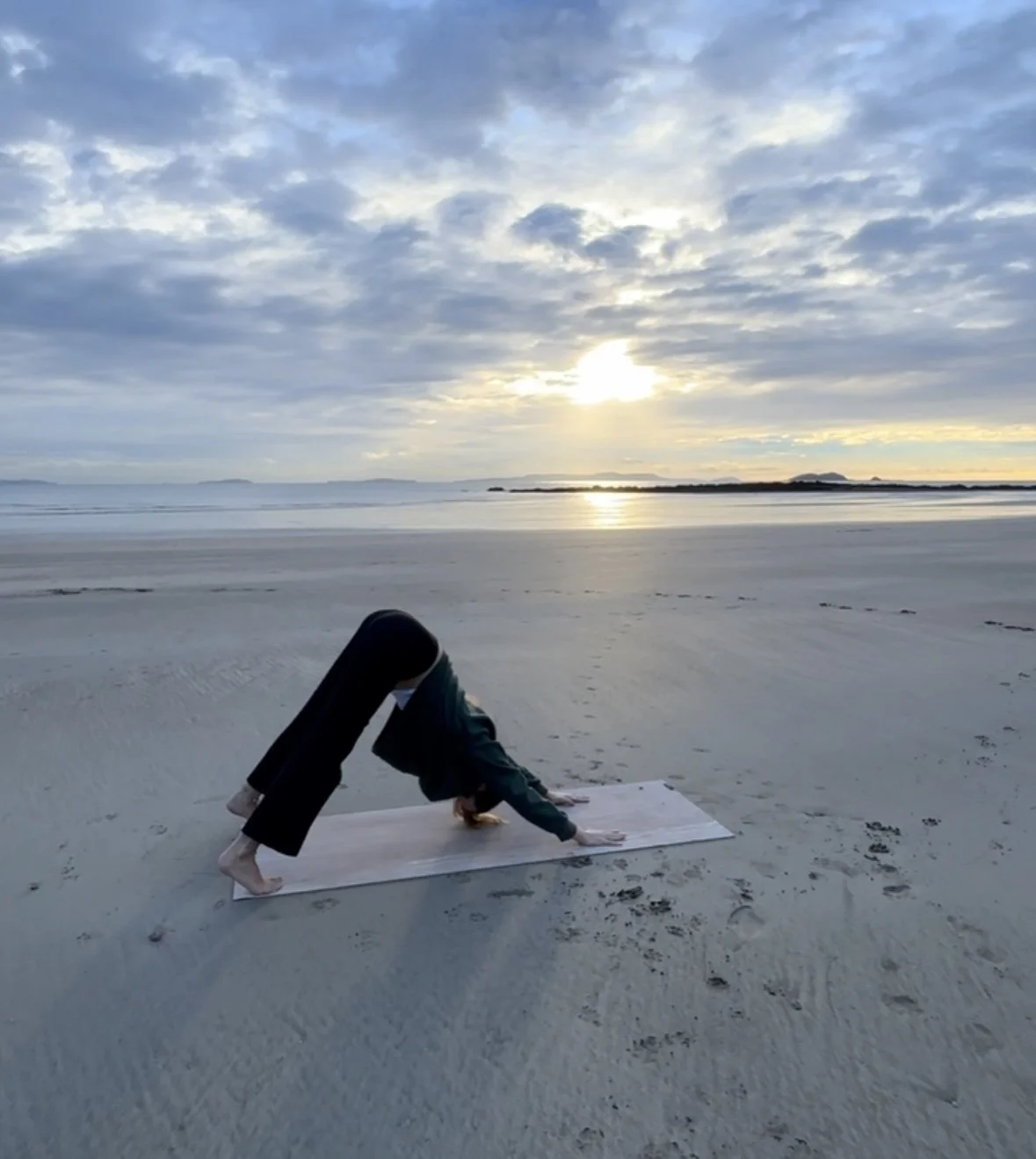Person practicing yoga on a mat on the beach at sunset with a cloudy sky and calm water in the background.