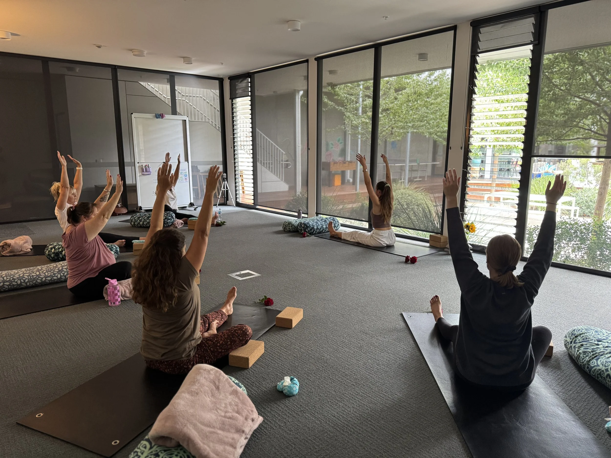 Group of people practicing yoga in a bright studio with large windows, some seated on mats and raising their arms, with the instructor leading at the front.