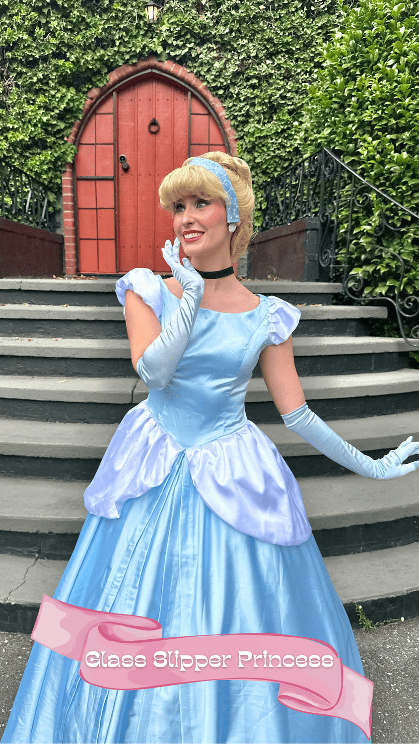 Woman dressed as a princess in a blue gown, standing on stairs outside with greenery and a red door in the background, holding her chin with one hand and smiling. A pink banner at the bottom reads 'Glass Slipper Princess'.