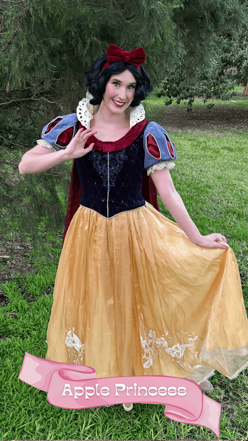 A woman dressed as the princess from the fairy tale Snow White, wearing a yellow skirt, blue and red top, with a red bow in her black hair, standing outdoors on grass with trees in the background.