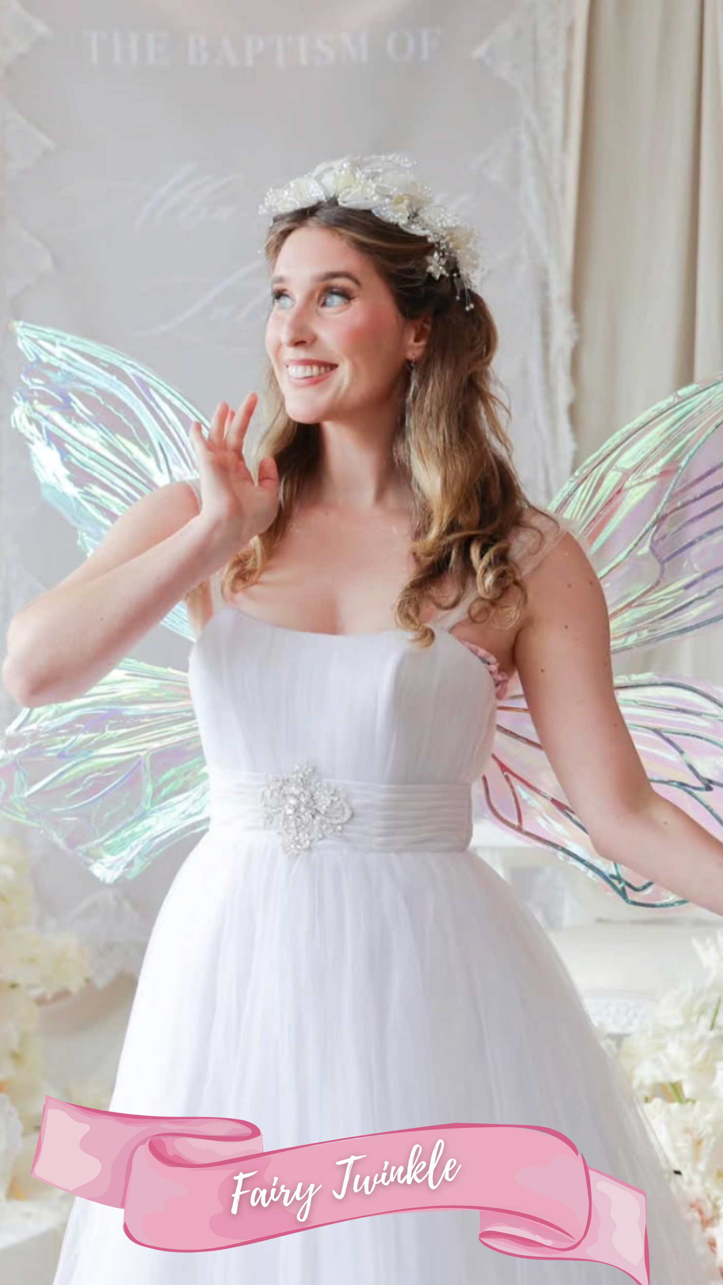 A young woman dressed as a fairy princess in a white dress with large white flowers and iridescent fairy wings, standing with a white floral backdrop.