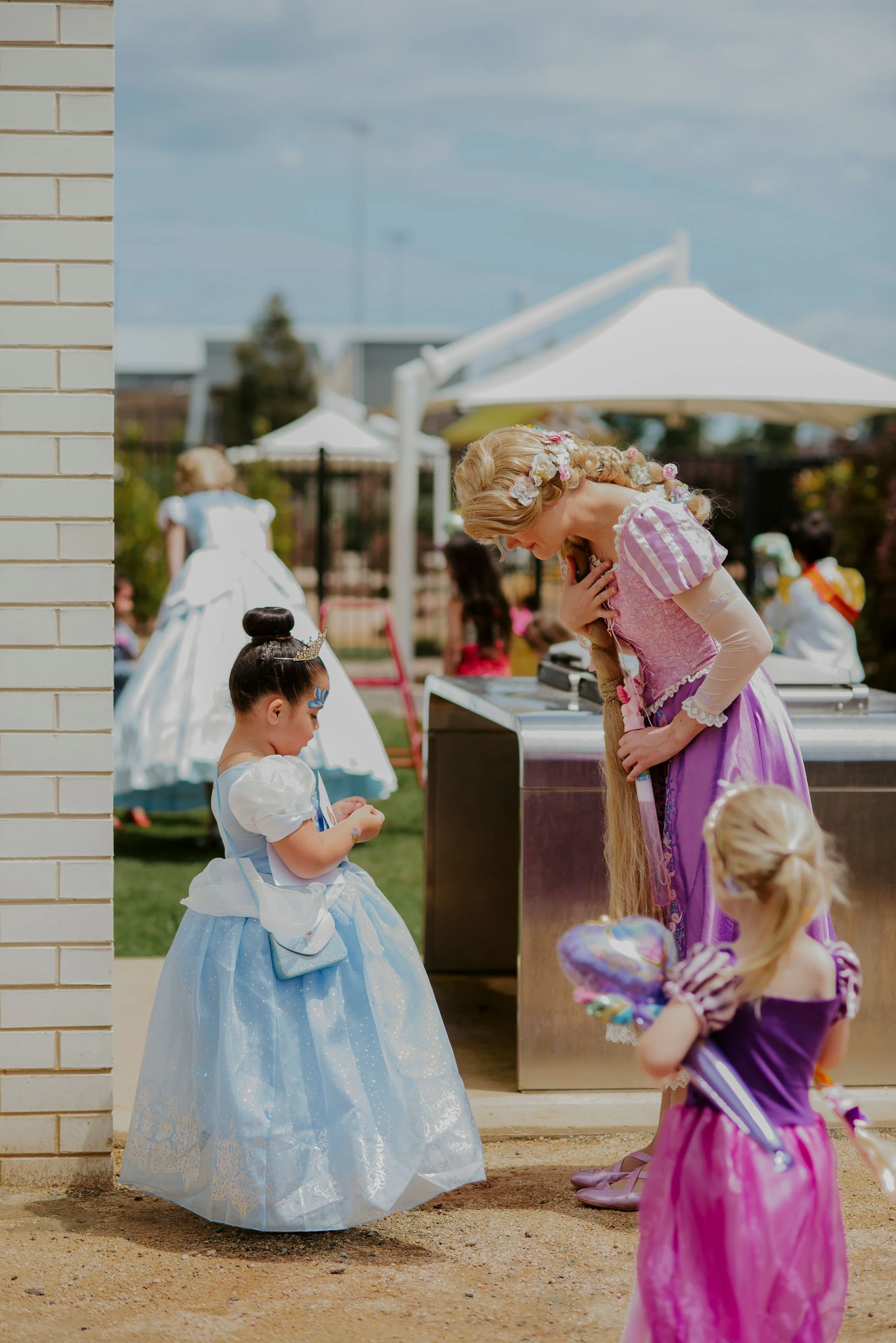 A woman dressed as Rapunzel with long braided hair and floral headband, interacting with young girls wearing princess costumes at a party outdoors. A girl in a blue dress with puffed sleeves and tiara is in front of her, and another girl with a purple dress and balloon is nearby. In the background, there are people, a white canopy, and a white dress-clad child at a distance.