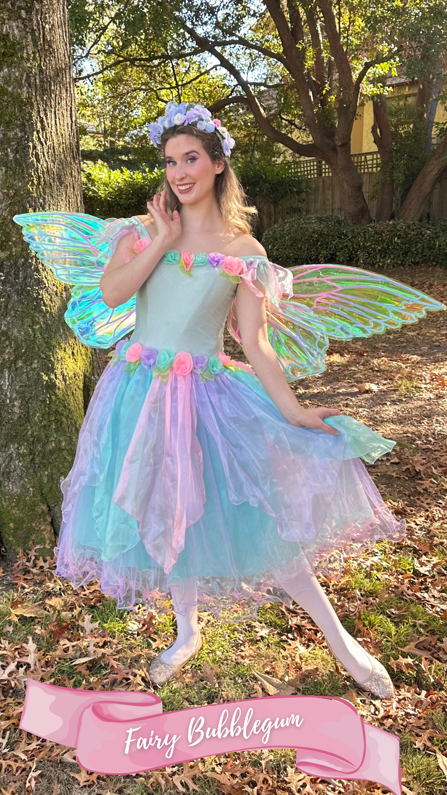 A woman dressed as a fairy with iridescent butterfly wings, wearing a pastel-colored dress with flower accents, a floral headpiece, standing outdoors in a wooded area during daytime.