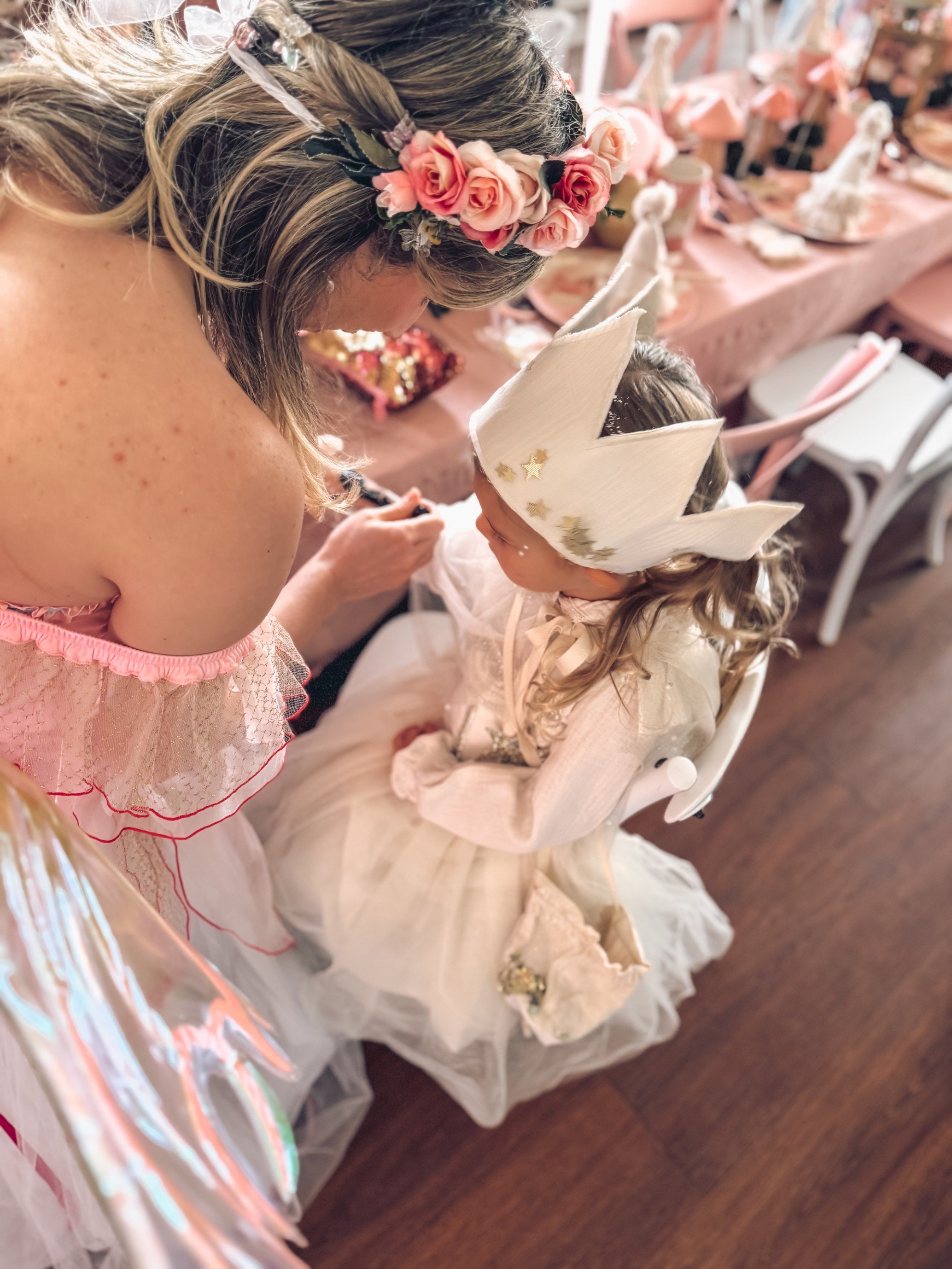 A woman with a floral headband is leaning towards a young girl dressed as a princess, who is sitting in a high chair. The girl is wearing a crown with star embellishments and a princess dress. The setting appears to be a birthday celebration with a decorated table in the background.