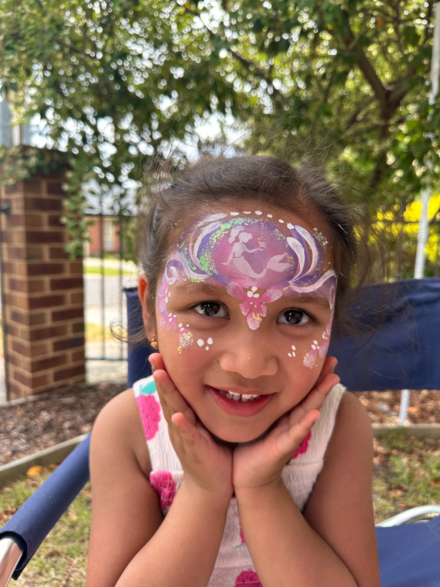 Young girl with face paint of a fairy surrounded by swirls, stars, and sparkles, outdoors with trees in the background.