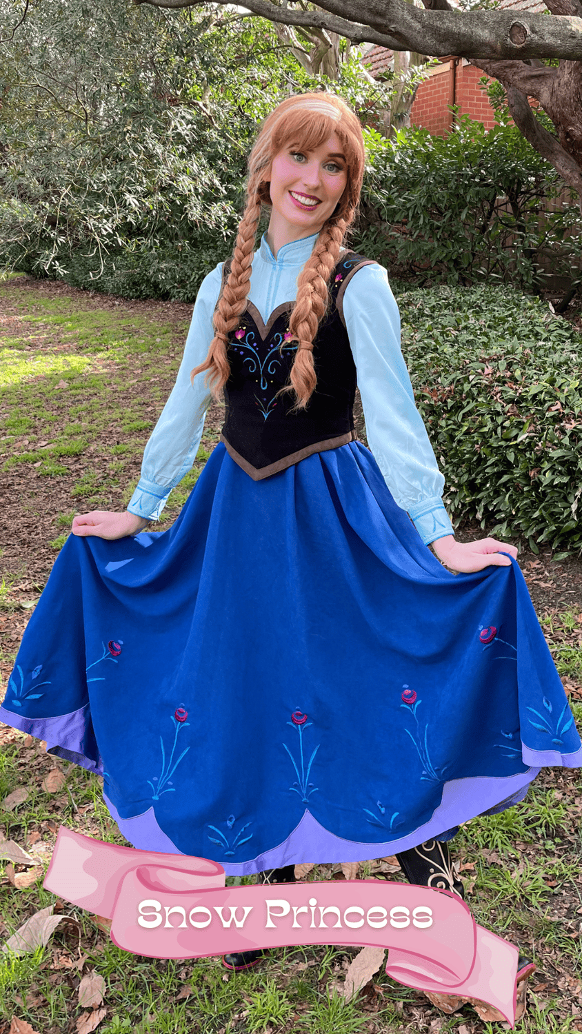 A woman dressed as a Snow Princess in a blue dress with embroidered floral details, standing outdoors in a garden with lush green foliage and trees, smiling at the camera.