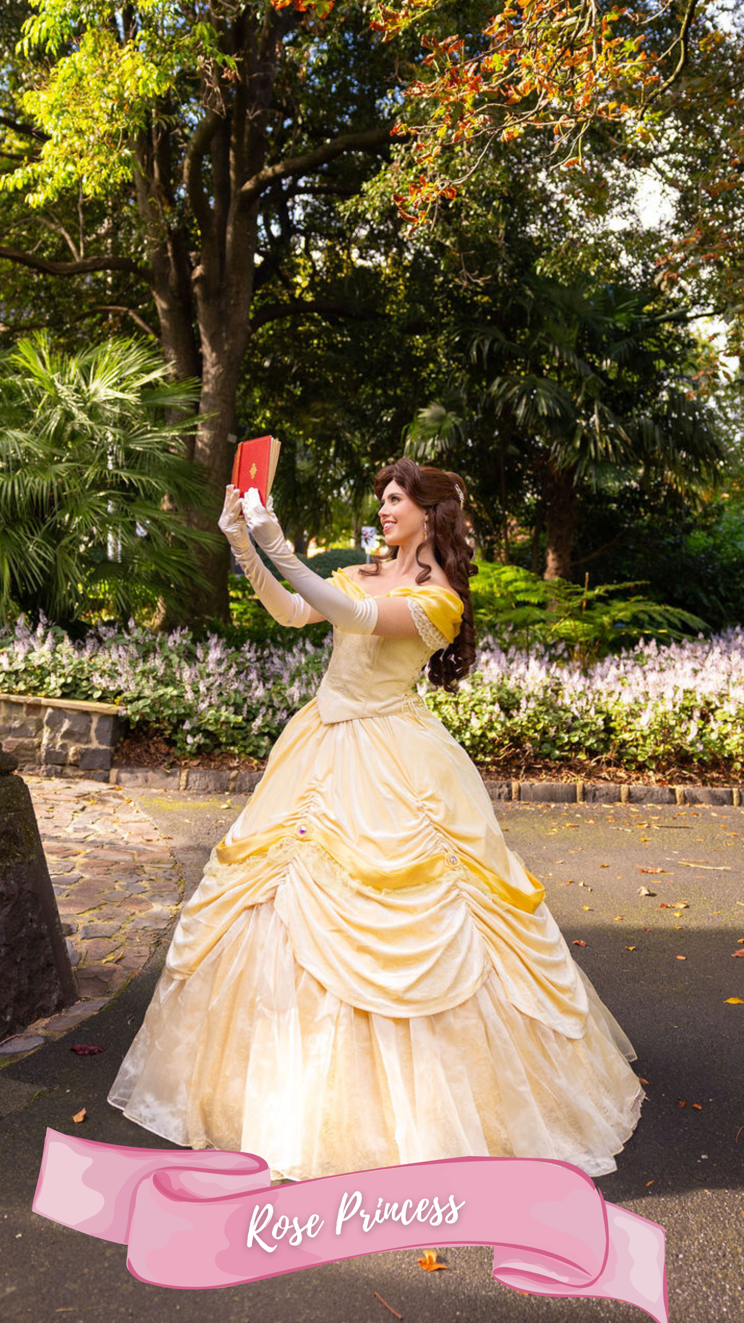 A woman dressed as a princess in a gold ball gown with a ruffled skirt and off-the-shoulder sleeves stands against a green hedge. She has long, curly auburn hair and wears long, matching gold gloves. She is smiling and has one hand on her chest, the 
