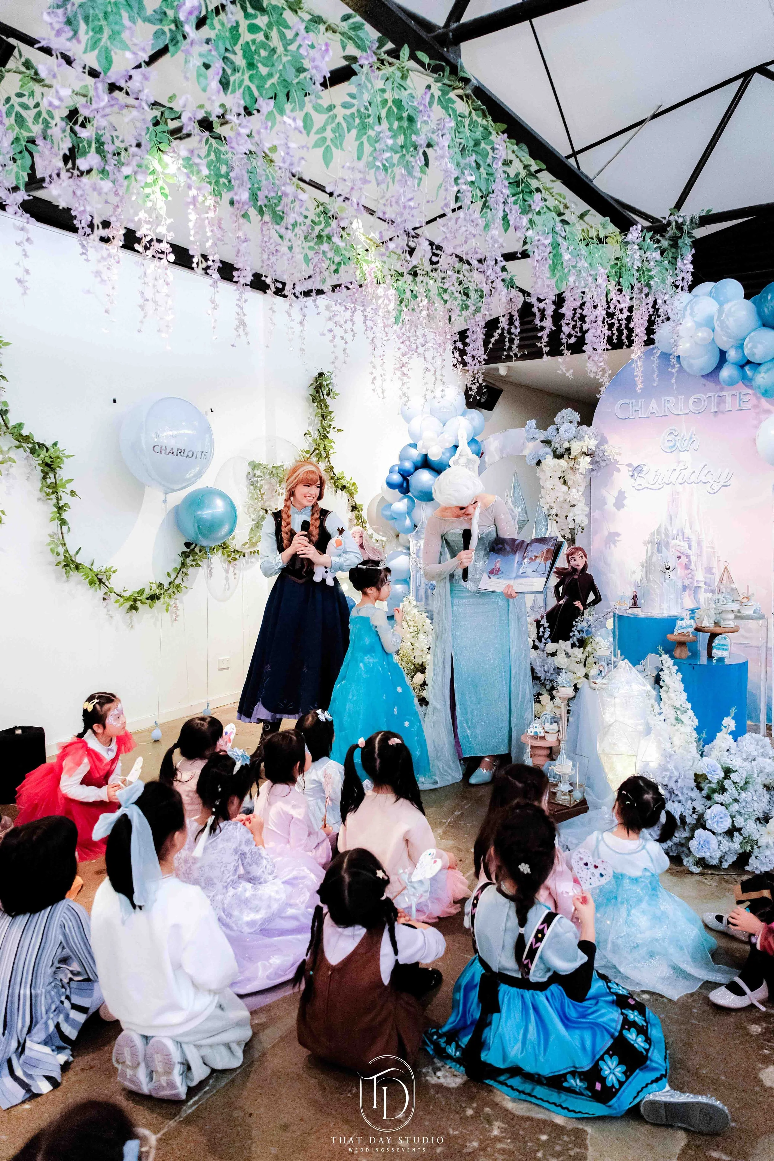Children sitting on the floor at a princess-themed birthday party with characters dressed as Elsa and Anna from Frozen, party decorations including balloons, flowers, and a backdrop with the name Charlotte and the number 6.