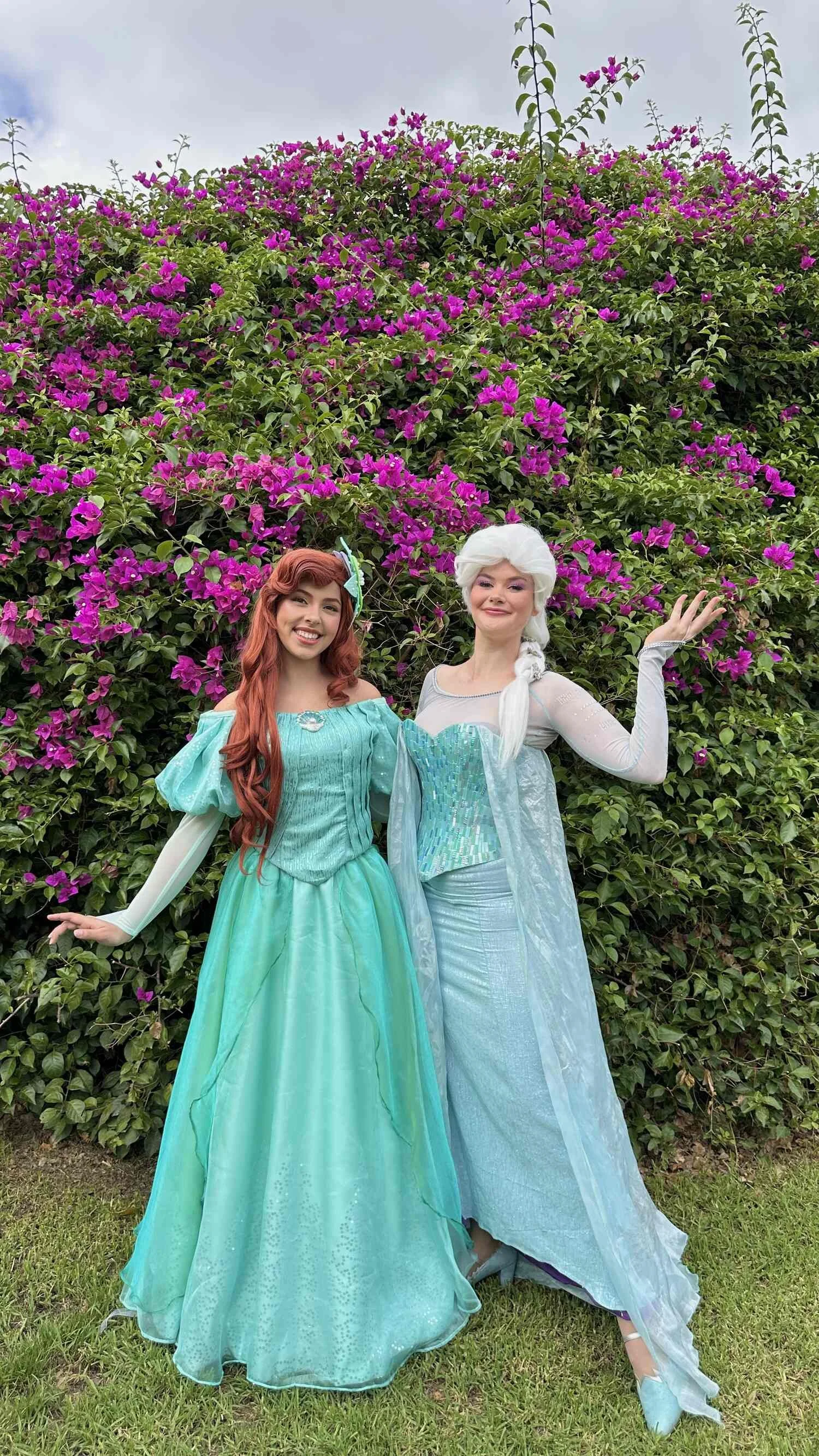 Two women dressed as Disney princesses, one with red hair in a turquoise gown and the other with white hair in a light blue gown, standing in front of a large bush with pink flowers.
