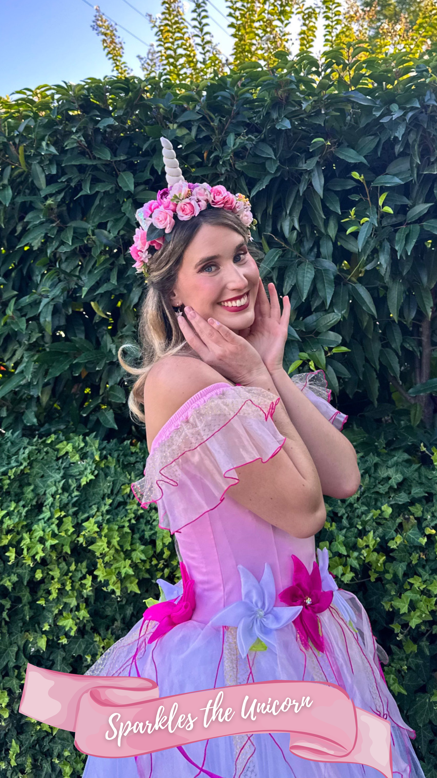 A young woman dressed as a unicorn princess, wearing a pink and white dress decorated with large pink and white flowers, standing outdoors in front of green foliage, with a floral unicorn horn headpiece on her head and smiling at the camera.