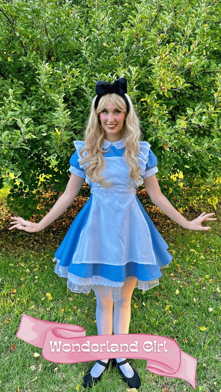 A woman dressed as Alice from Alice in Wonderland, wearing a blue dress with puffed sleeves, a white apron, and a black bow headband, standing outdoors in front of green trees, with a pink banner at her feet that reads 'Wonderland Girl'.