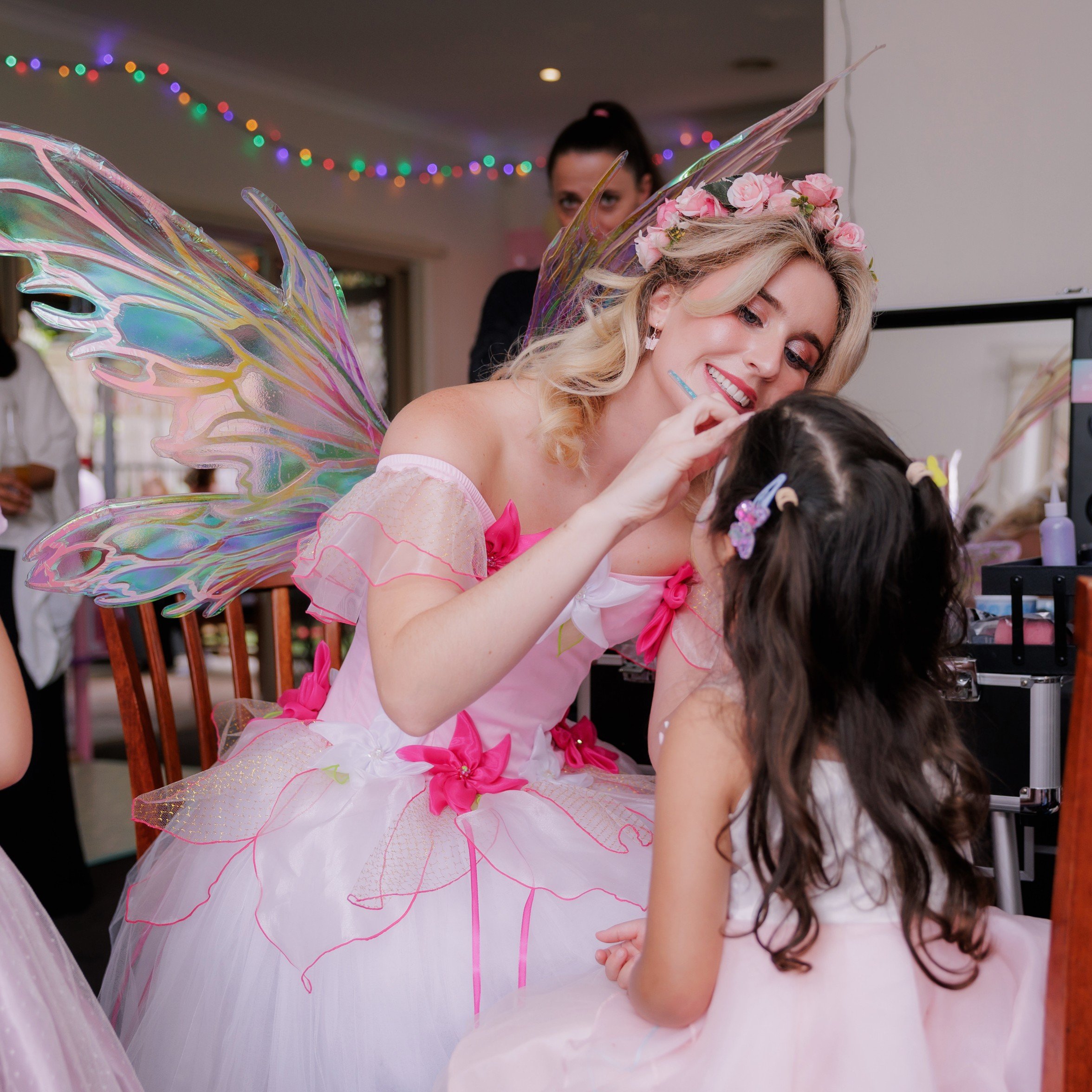 A fairy costume performer with iridescent fairy wings, a flower crown, and pink and white dress, applying makeup to a young girl during a celebration.