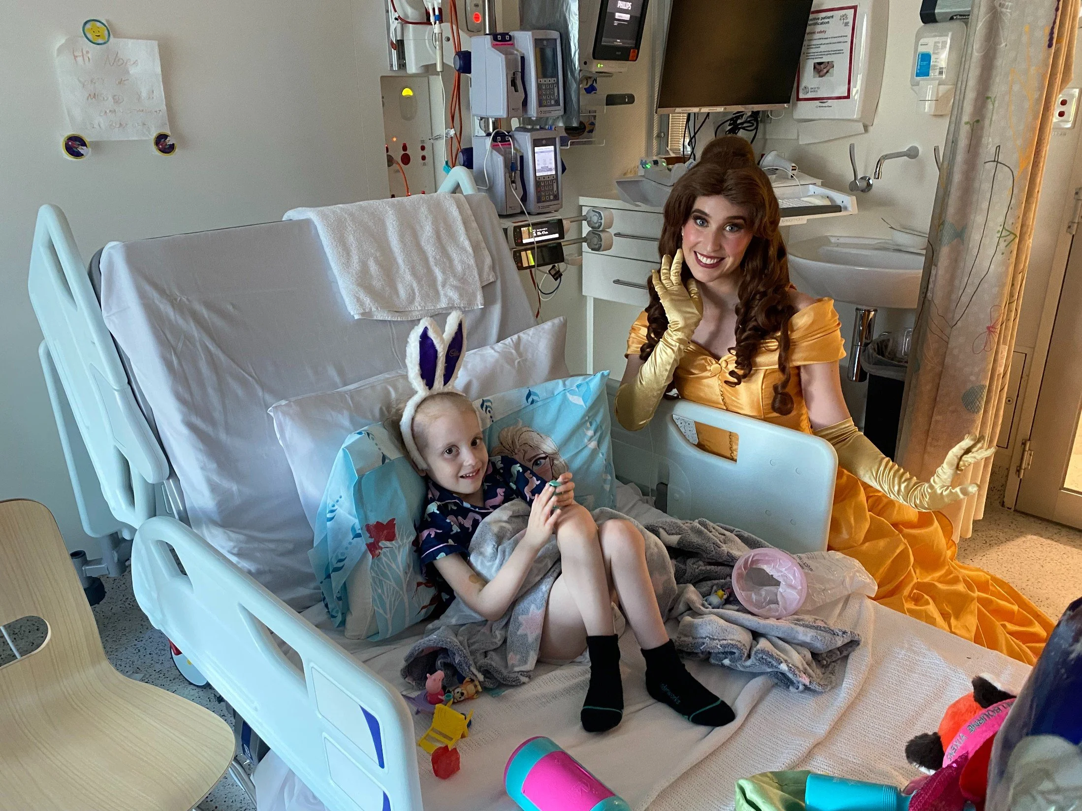 A young girl with bunny ears on her head sitting on a hospital bed in a colorful hospital room, next to a woman dressed as Belle from Disney's Beauty and the Beast in a golden dress with long gloves, posing happily.