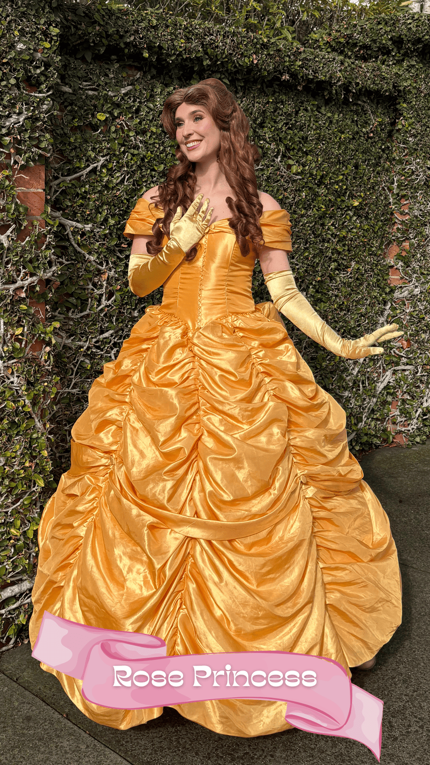 A woman dressed as a princess in a gold ball gown with a ruffled skirt and off-the-shoulder sleeves stands against a green hedge. She has long, curly auburn hair and wears long, matching gold gloves. She is smiling and has one hand on her chest, the 