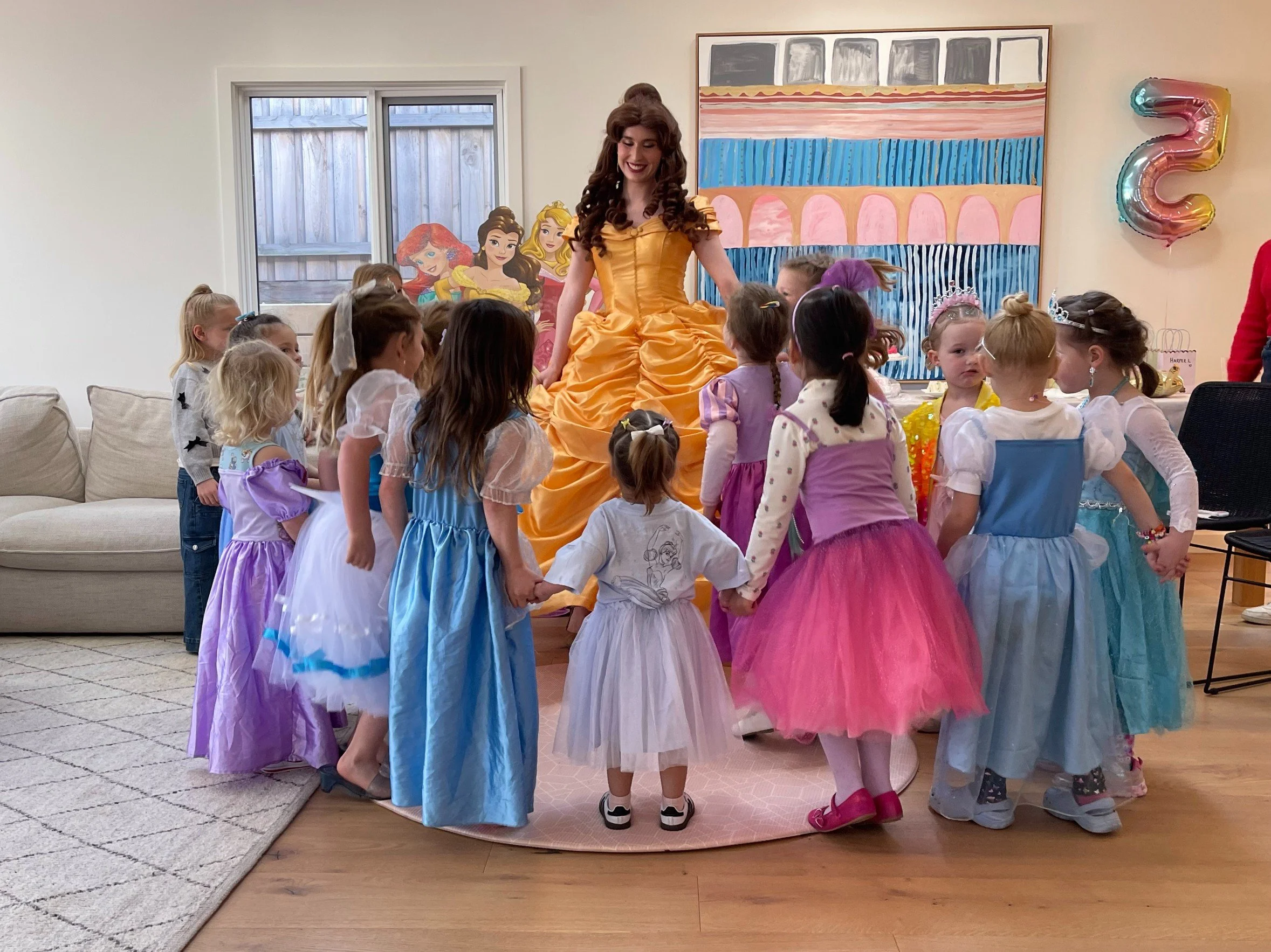 A woman dressed as a princess in a gold gown surrounded by young children at a birthday party in a decorated room.
