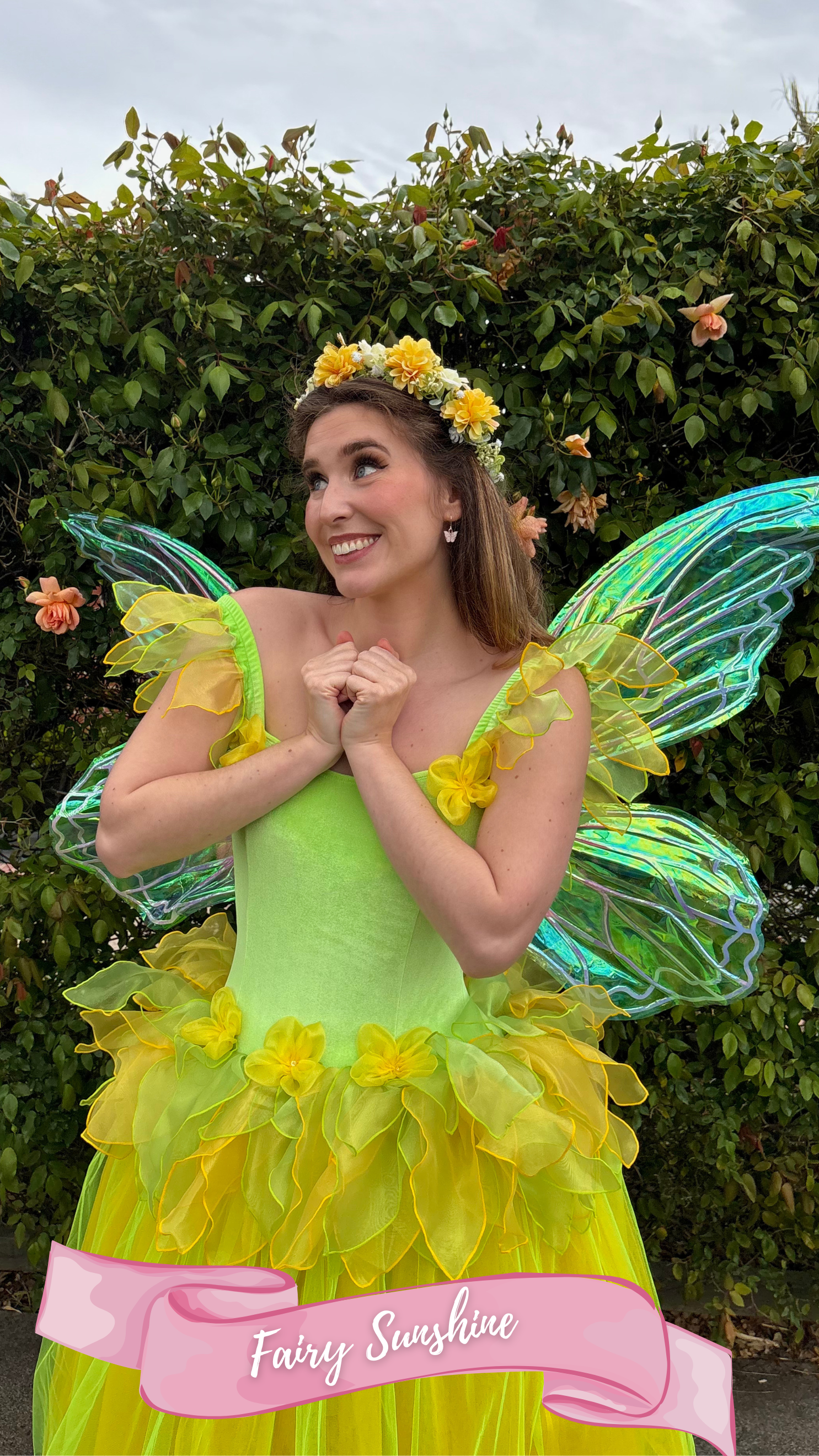 Woman dressed as a fairy fairy in a bright yellow and green costume with fairy wings, standing in front of a green leafy background and smiling.