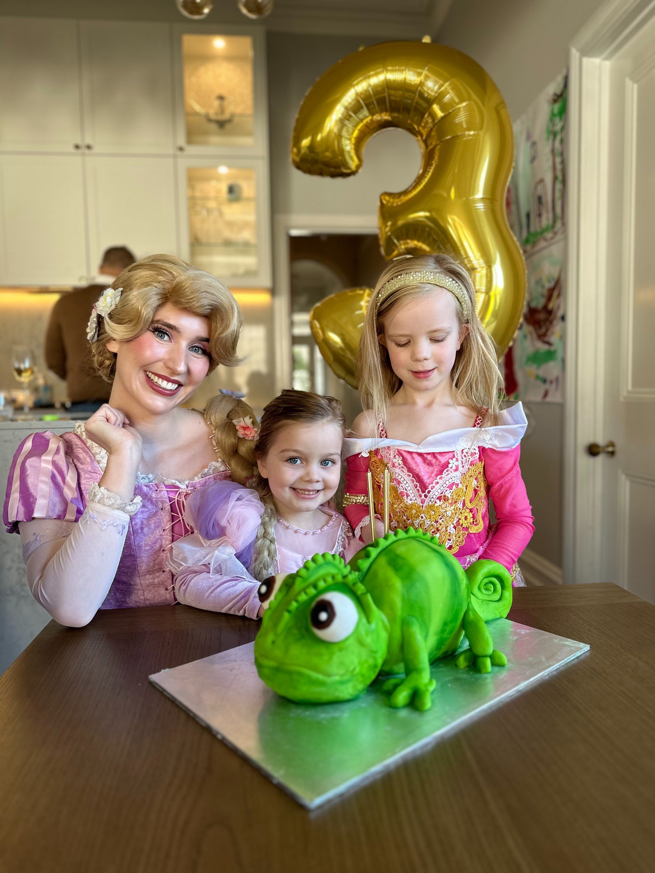 Three young girls and an adult woman, dressed in princess costumes, celebrating a birthday with a dinosaur-shaped cake in front of them. A large gold number 3 balloon is in the background.