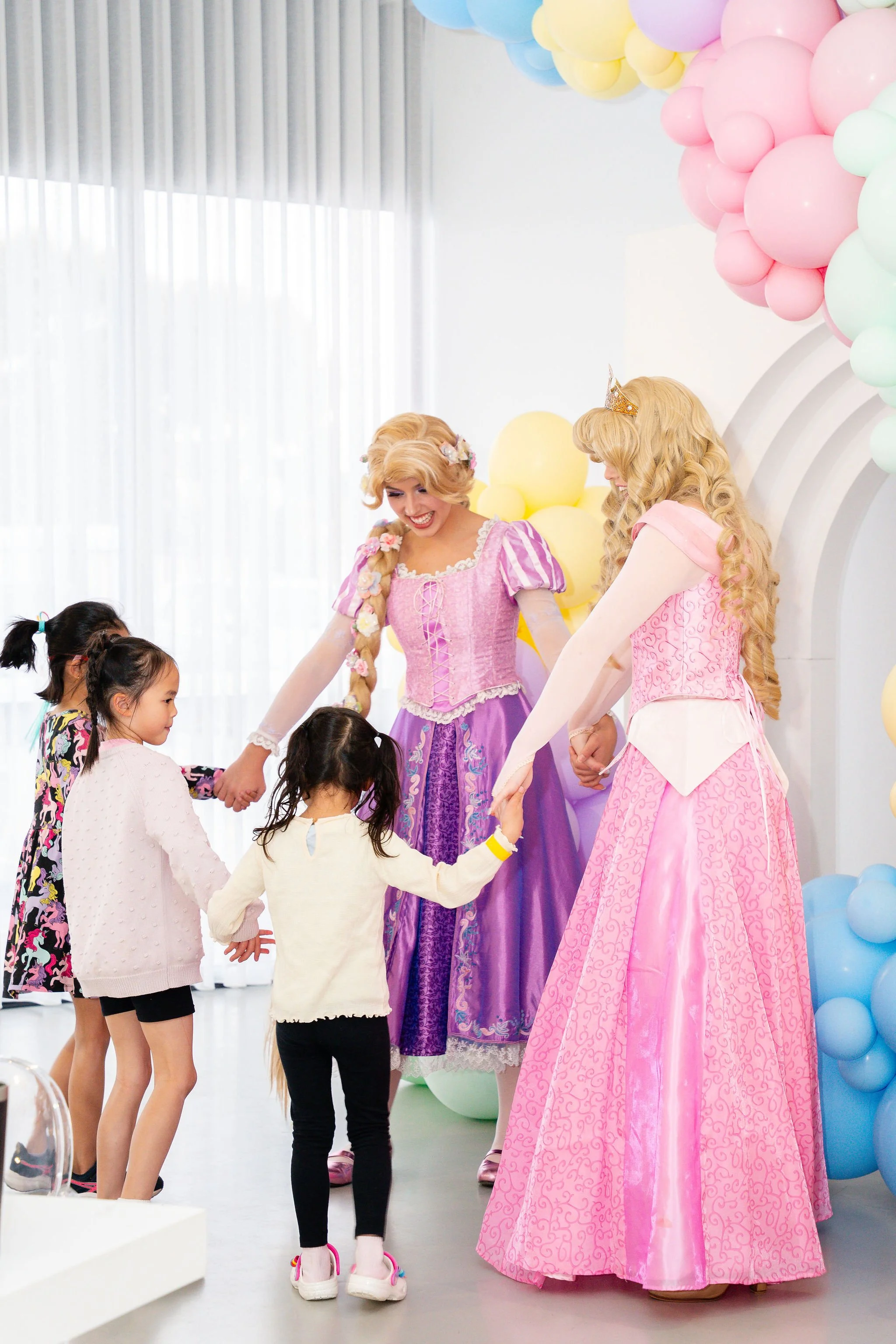 Children and two women dressed as princesses holding hands in a circle at a birthday party decorated with pastel-colored balloons.