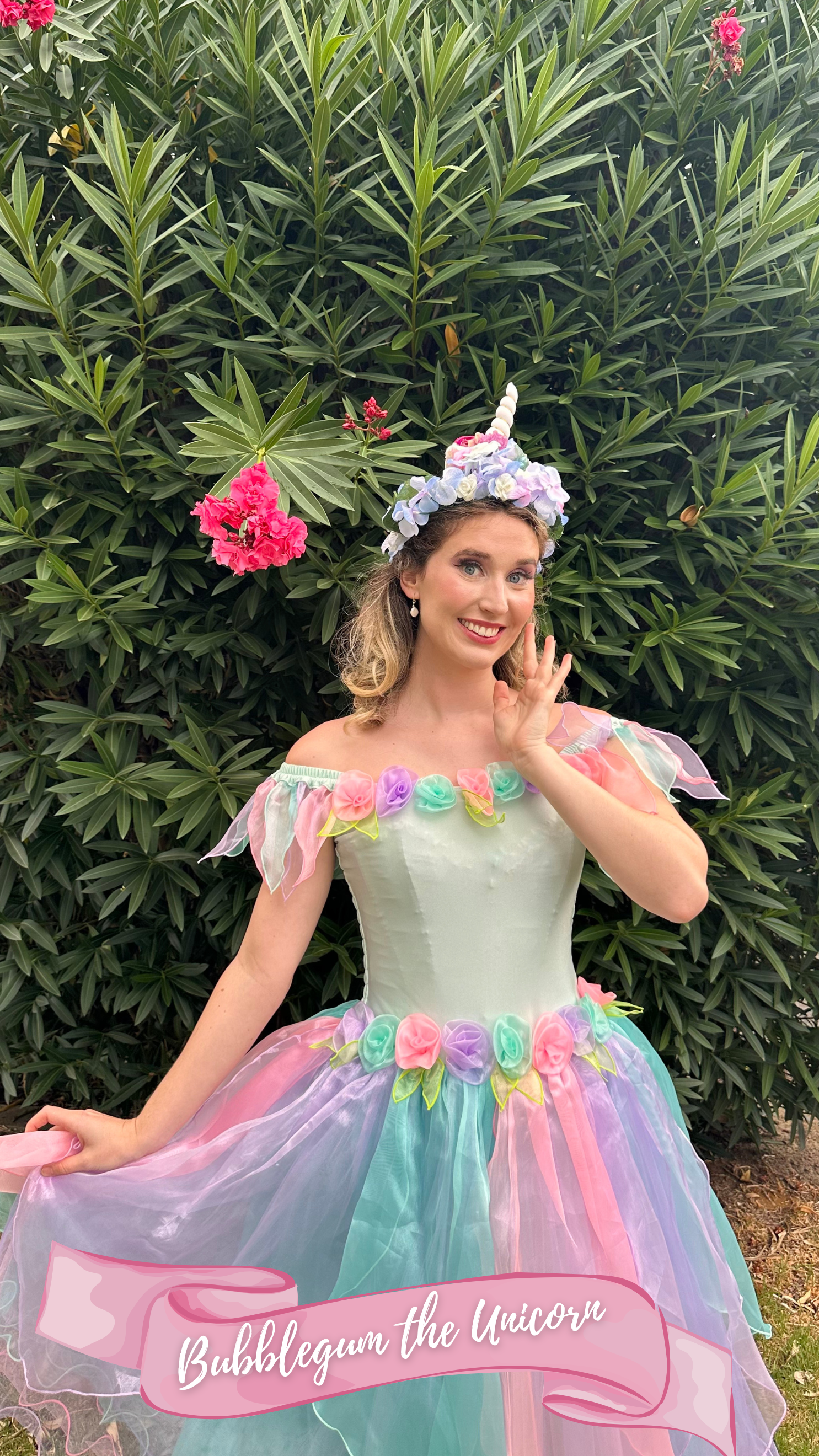 A young woman dressed as a unicorn princess, wearing a pastel-colored dress with flower accents, a floral headpiece, standing 
outdoors in front of green foliage, with a floral unicorn horn headpiece on her head and smiling at the camera.