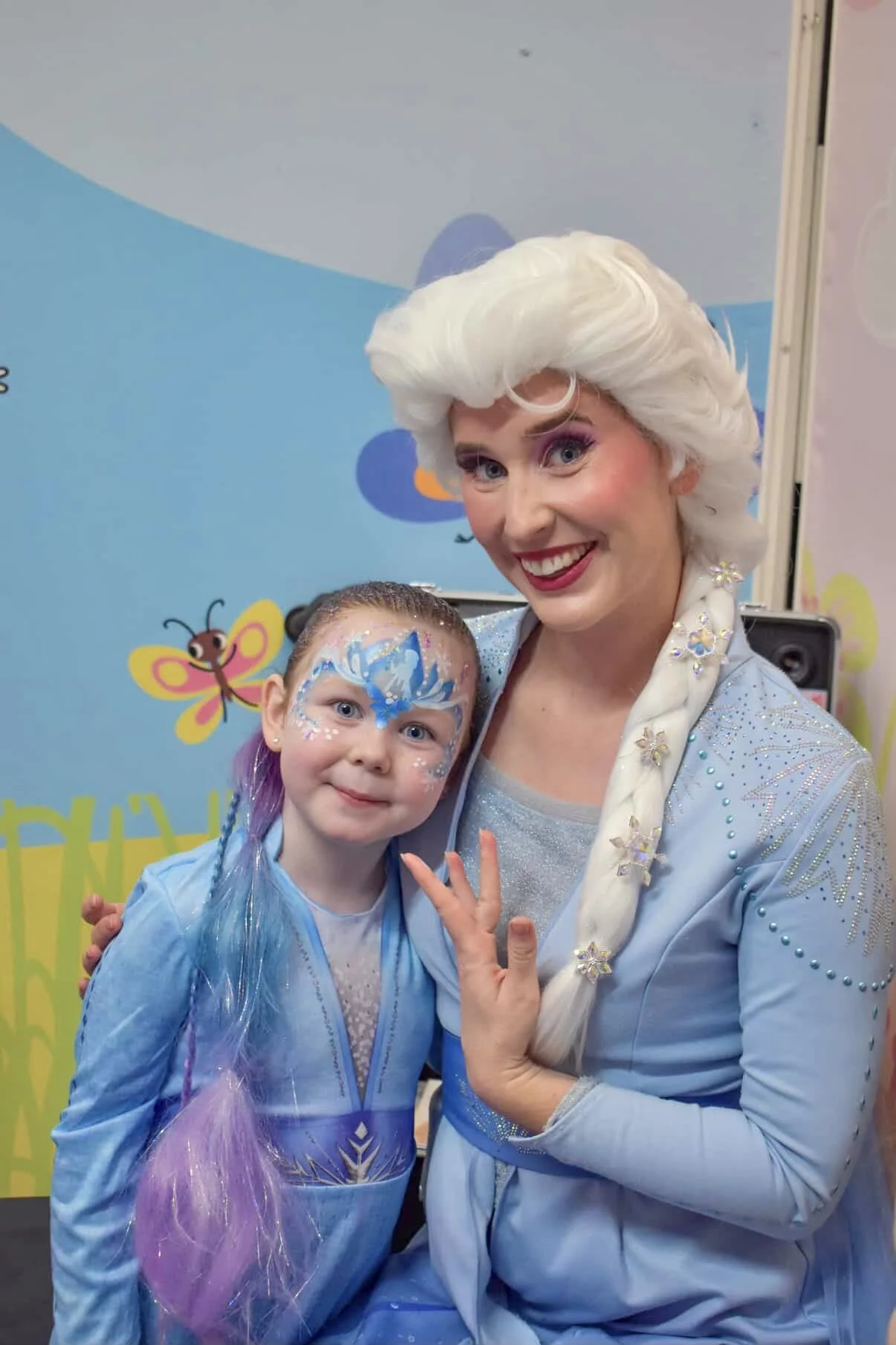 A woman dressed as Elsa from Frozen with white hair and a blue costume, smiling and posing with a young girl who has face paint resembling Elsa's face, with blue and white colors and a butterfly design, in front of a blue background with cartoon bees and a butterfly.