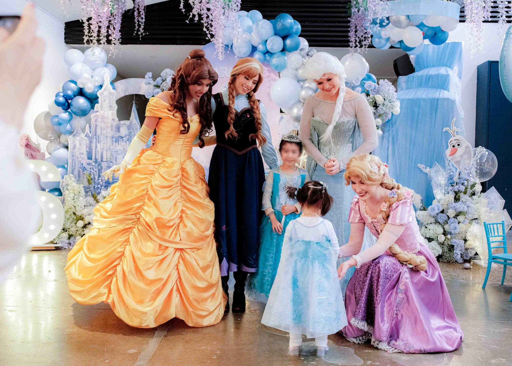 Disney princess characters, including Belle, Anna, Elsa, and Rapunzel, meet young children at a themed party with blue, white, and purple decorations and balloons.
