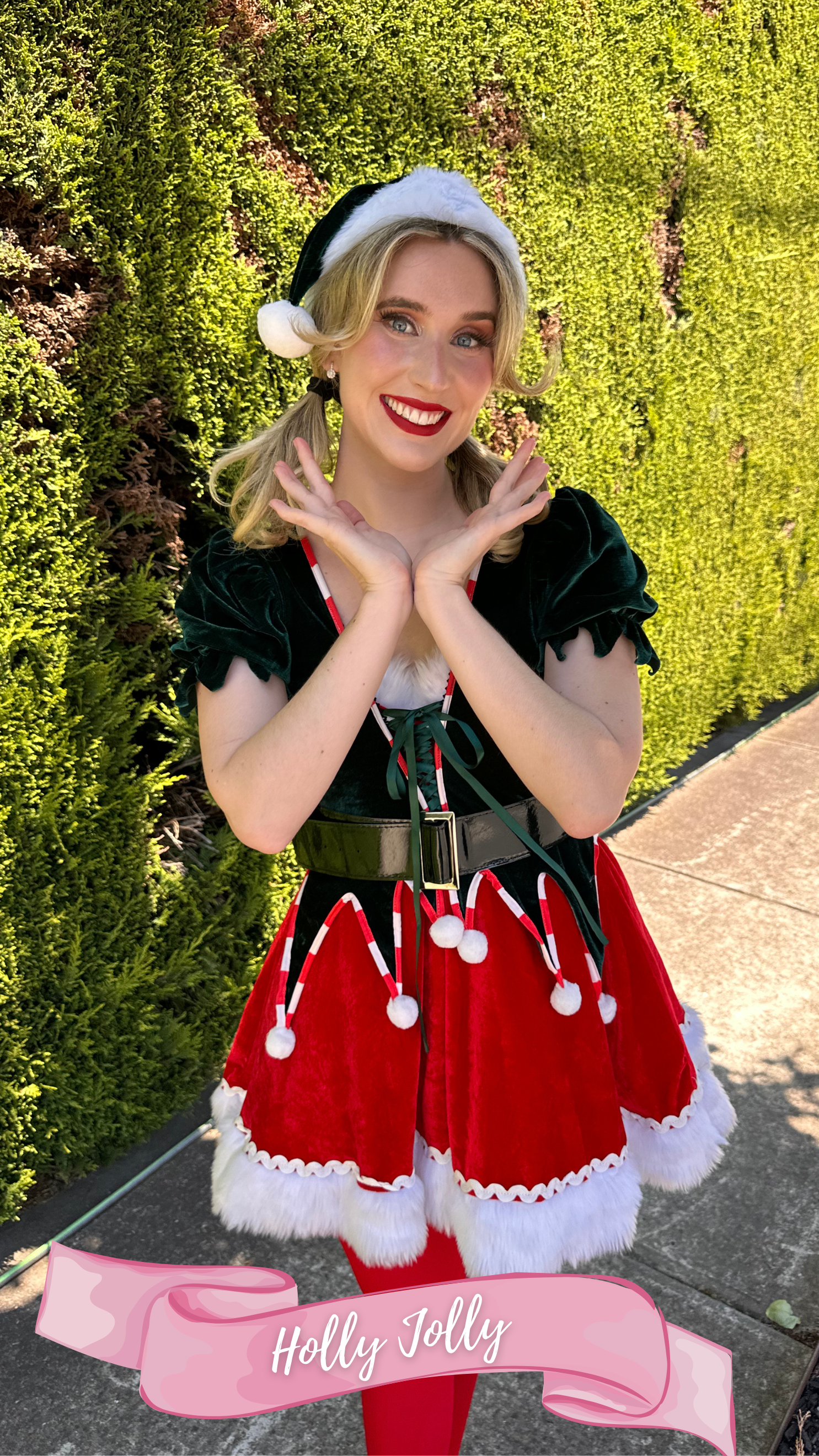 A young woman smiling in a festive holiday outfit with a Santa hat, green velvet top, red skirt with white trimmings, red tights, standing outdoors in front of a green hedge. The bottom of the image features a pink banner with the text 'Holly Jolly t