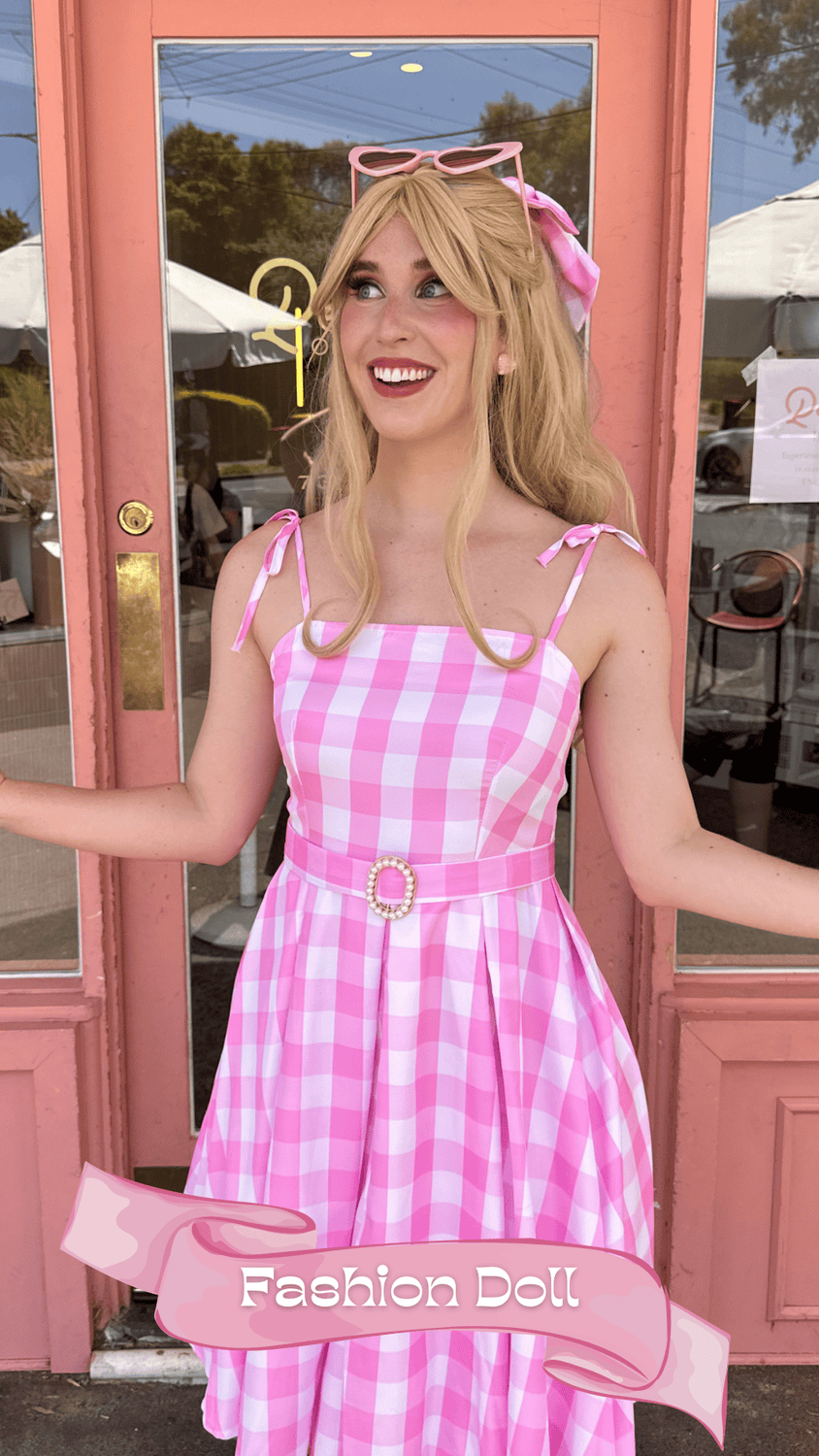 A woman standing in front of a pink building, wearing a pink and white checkered dress with shoulder ties, a matching pink belt with a pearl buckle, pink sunglasses on her head, and a pink hair scarf, smiling cheerfully.