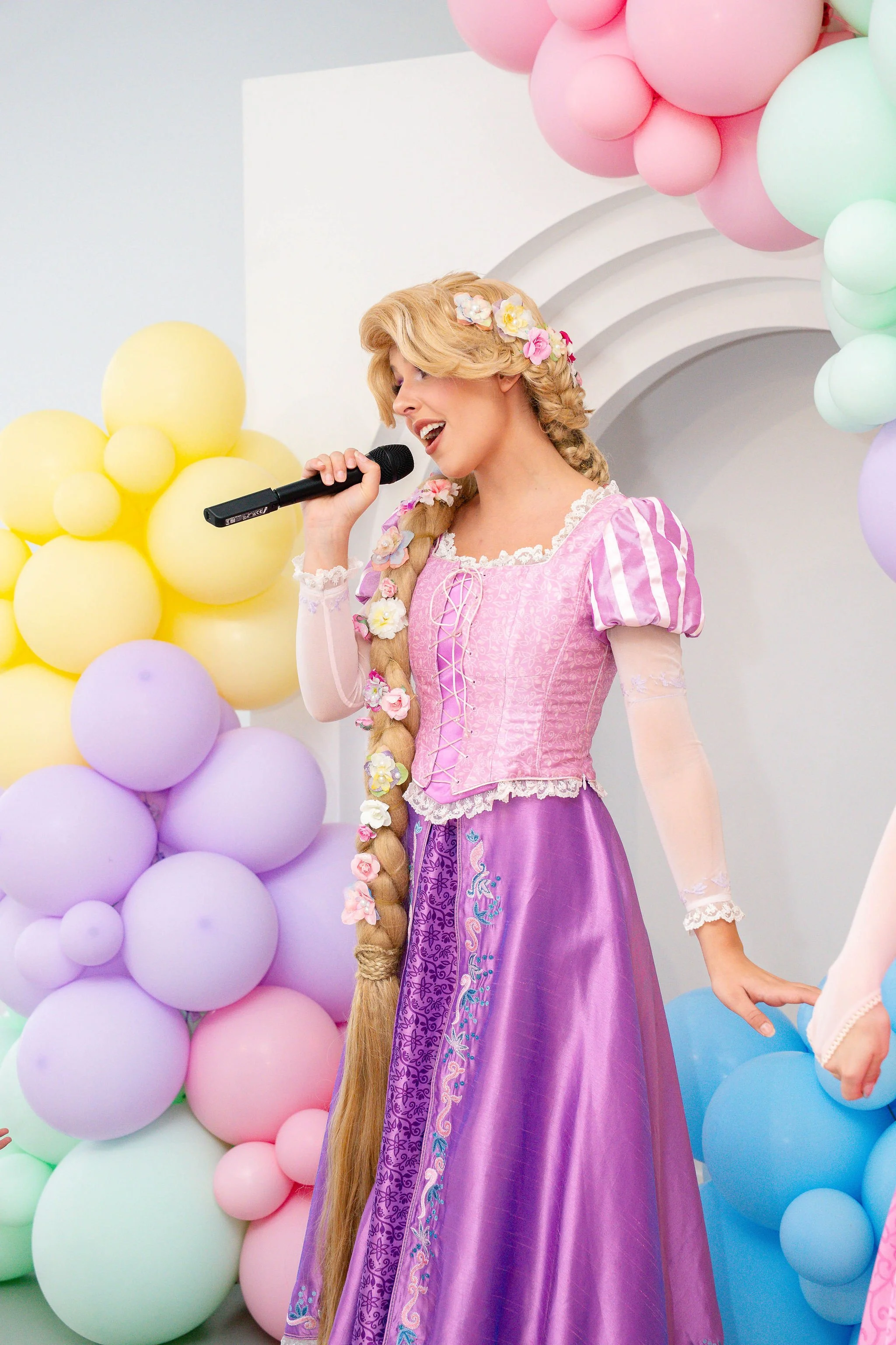 A woman dressed as Rapunzel in a pink and purple costume, singing into a microphone at a colorful balloon party.