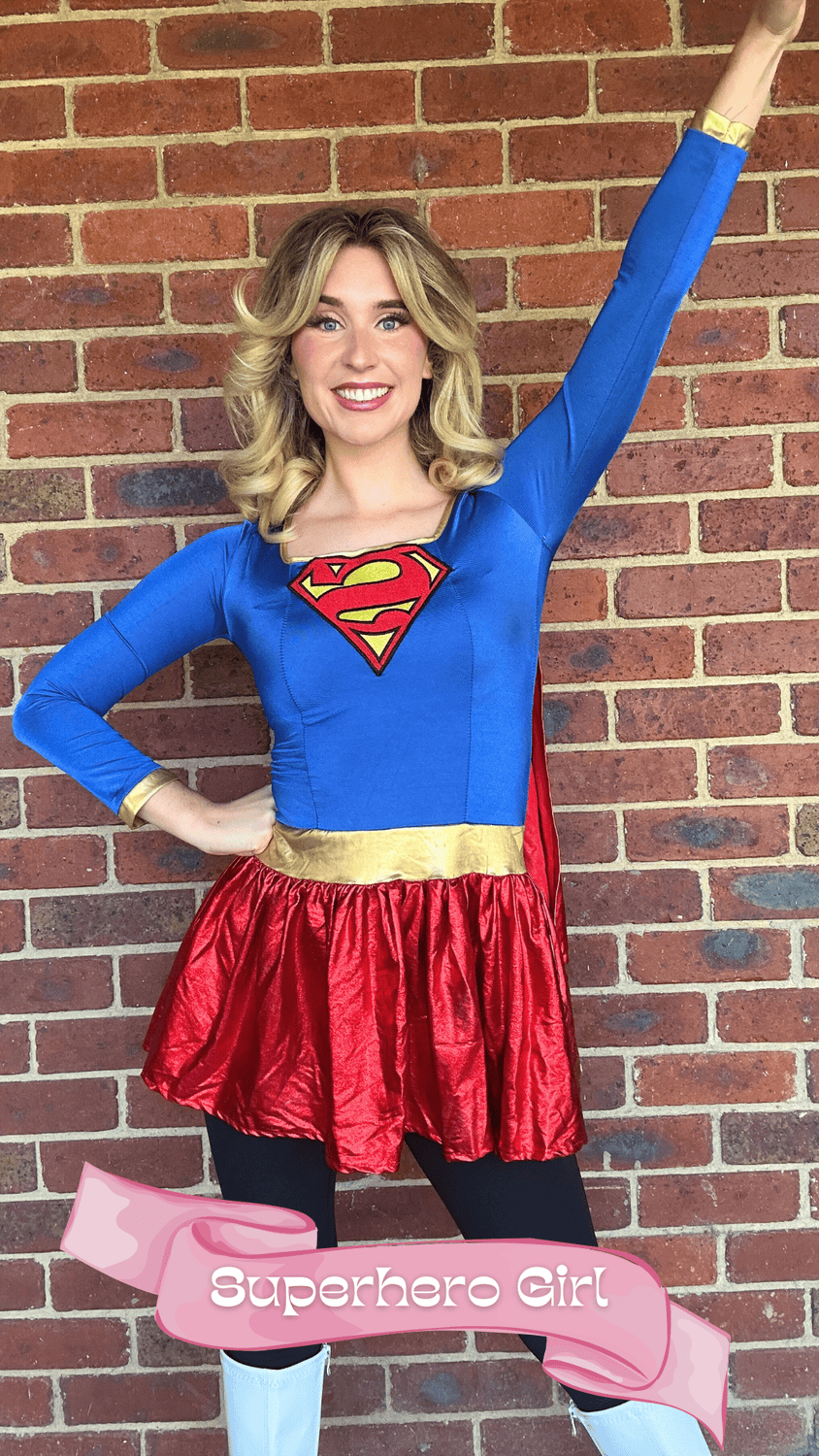 Young woman dressed as a superhero girl in a blue and red costume with a 'Superman' logo on her chest, posing confidently with one arm raised and smiling against a brick wall background. A pink banner at the bottom reads 'Superhero Girl'.