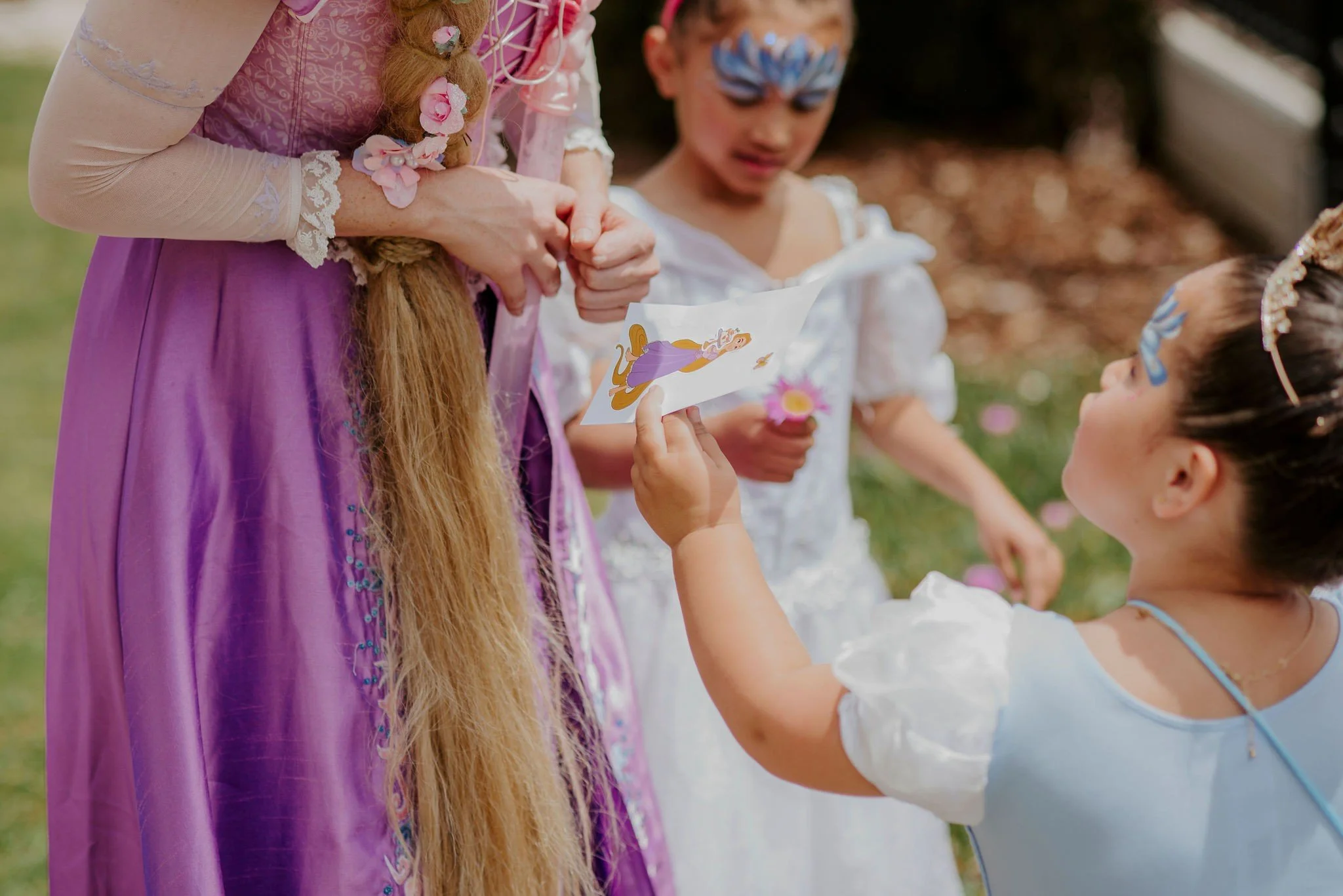Two young girls and an adult woman dressed as princesses during a fairy tale-themed event outside, with one girl holding a storybook and the other girl looking at her.
