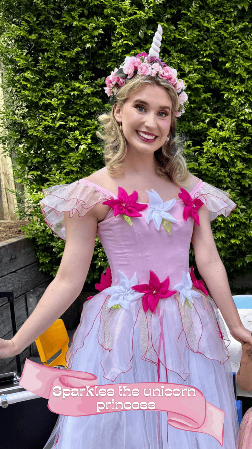 A young woman dressed as a unicorn princess, wearing a pink and white dress decorated with large pink and white flowers, standing outdoors in front of green foliage, with a floral unicorn horn headpiece on her head and smiling at the camera.