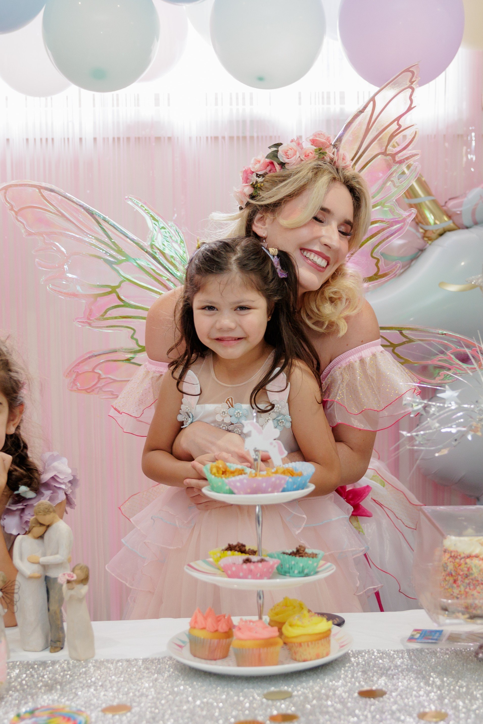 A woman dressed as a fairy with pink wings and a floral crown is hugging a young girl with butterfly wings and a dress, at a birthday party with cupcakes and decorations.