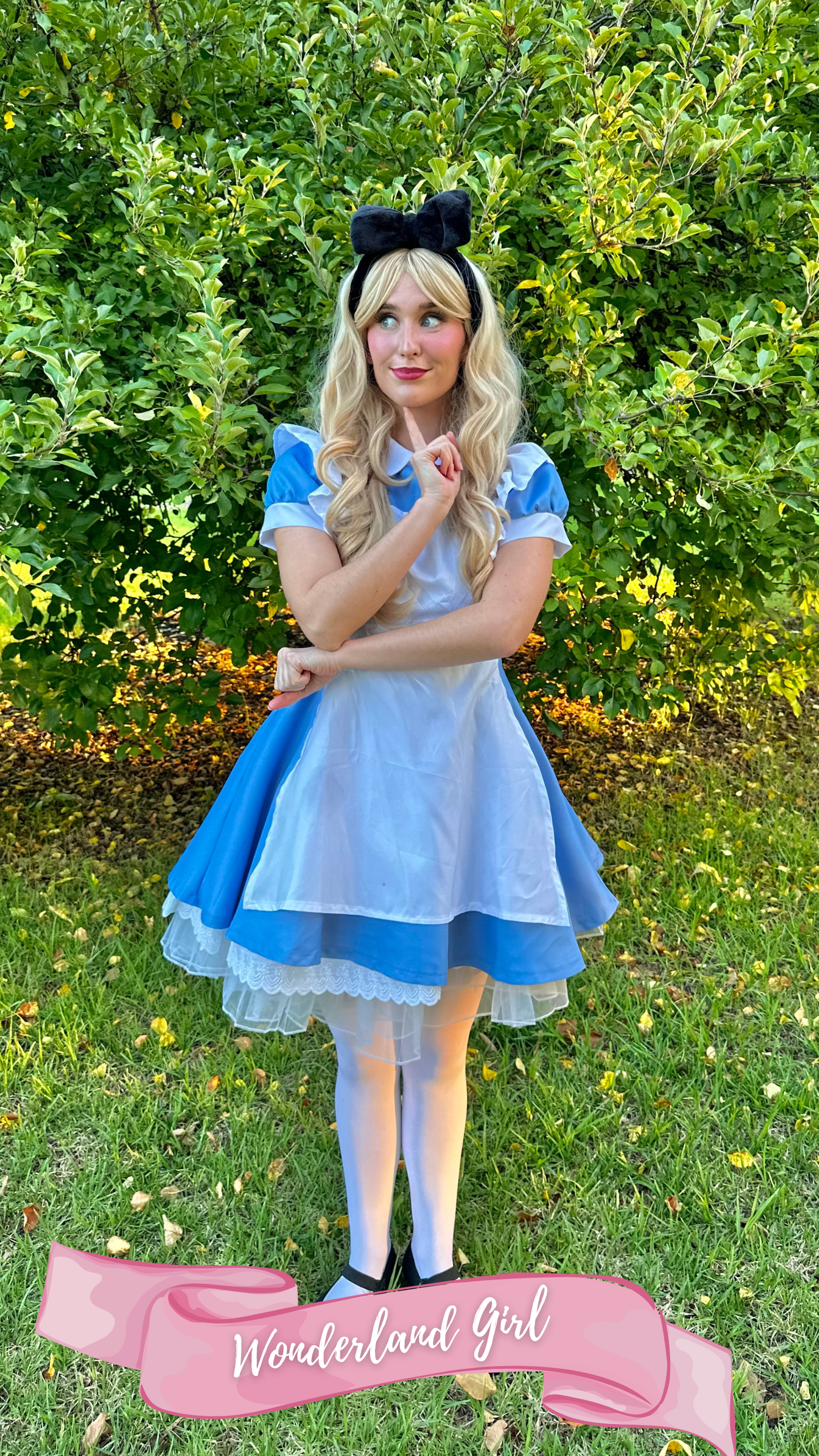 A woman dressed as Alice from Alice in Wonderland, wearing a blue dress with puffed sleeves, a white apron, and a black bow headband, standing outdoors in front of green trees, with a pink banner at her feet that reads 'Wonderland Girl'.