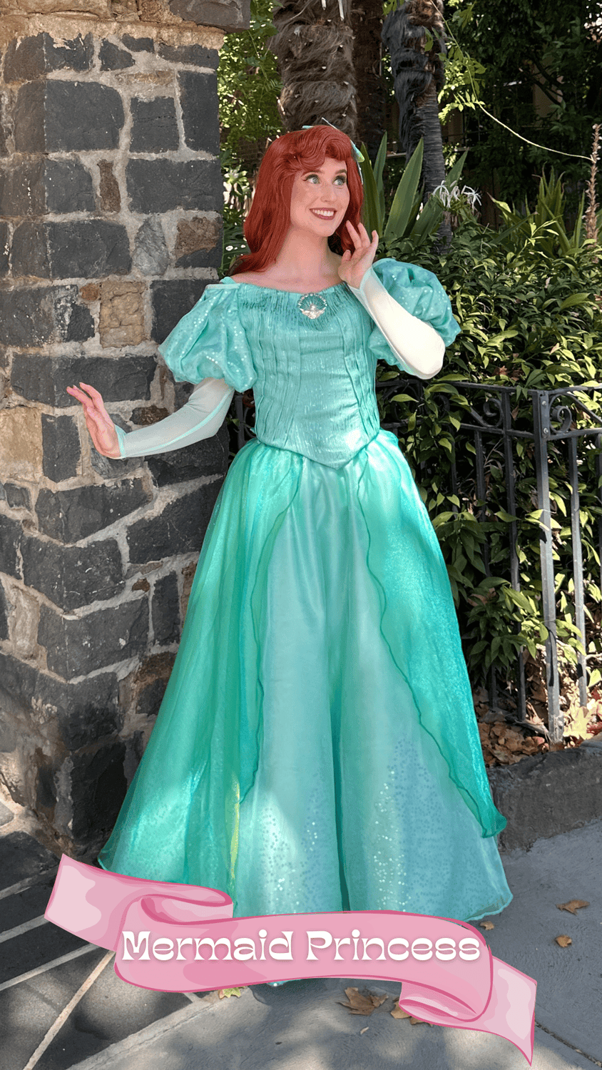 A woman dressed as a mermaid princess with red hair, wearing a shiny turquoise gown with puffed sleeves, standing outdoors near a brick wall and greenery, with a pink banner at the bottom that says 'Mermaid Princess'.