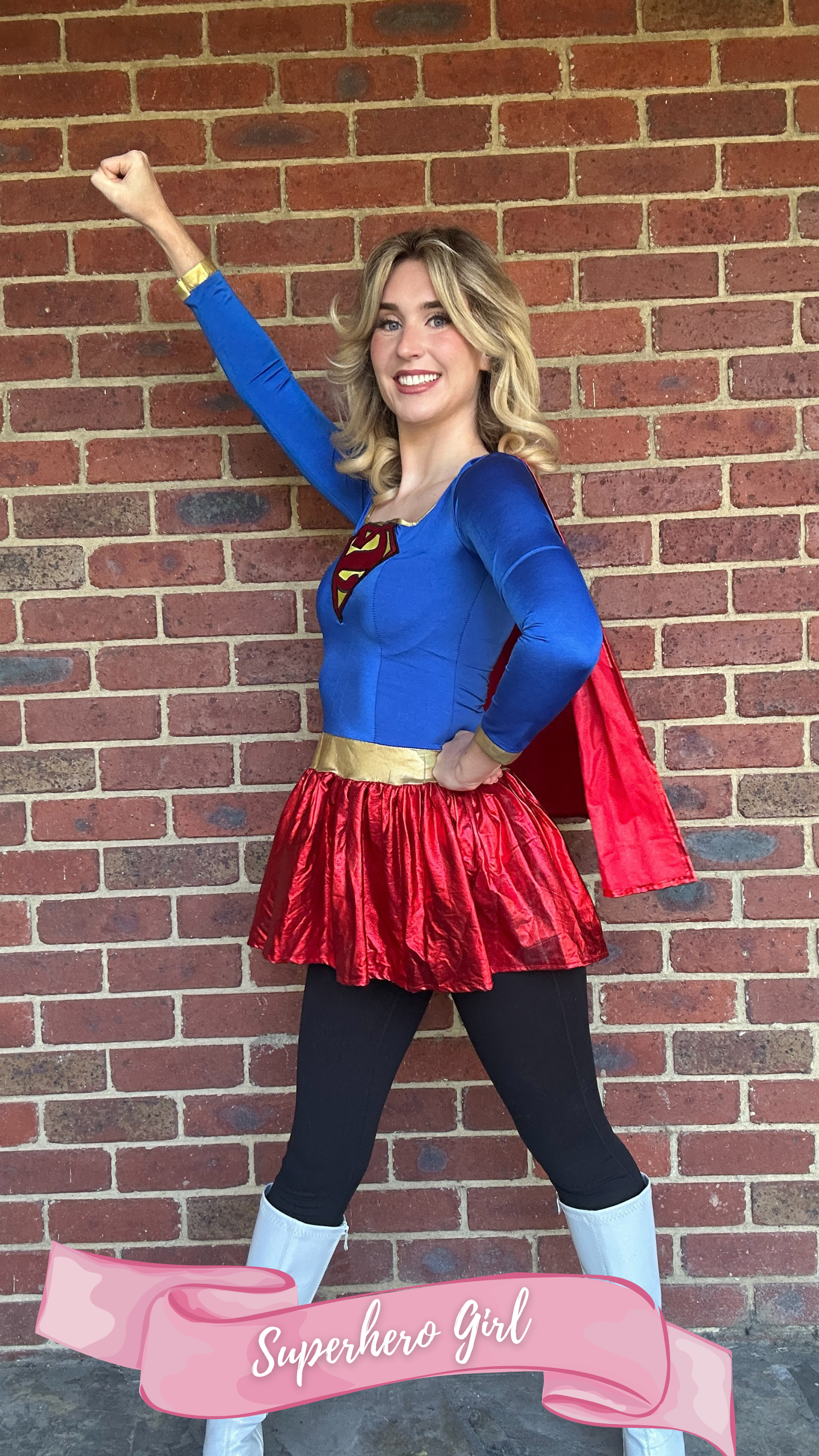 Young woman dressed as a superhero girl in a blue and red costume with a 'Superman' logo on her chest, posing confidently with one arm raised and smiling against a brick wall background. A pink banner at the bottom reads 'Superhero Girl'.