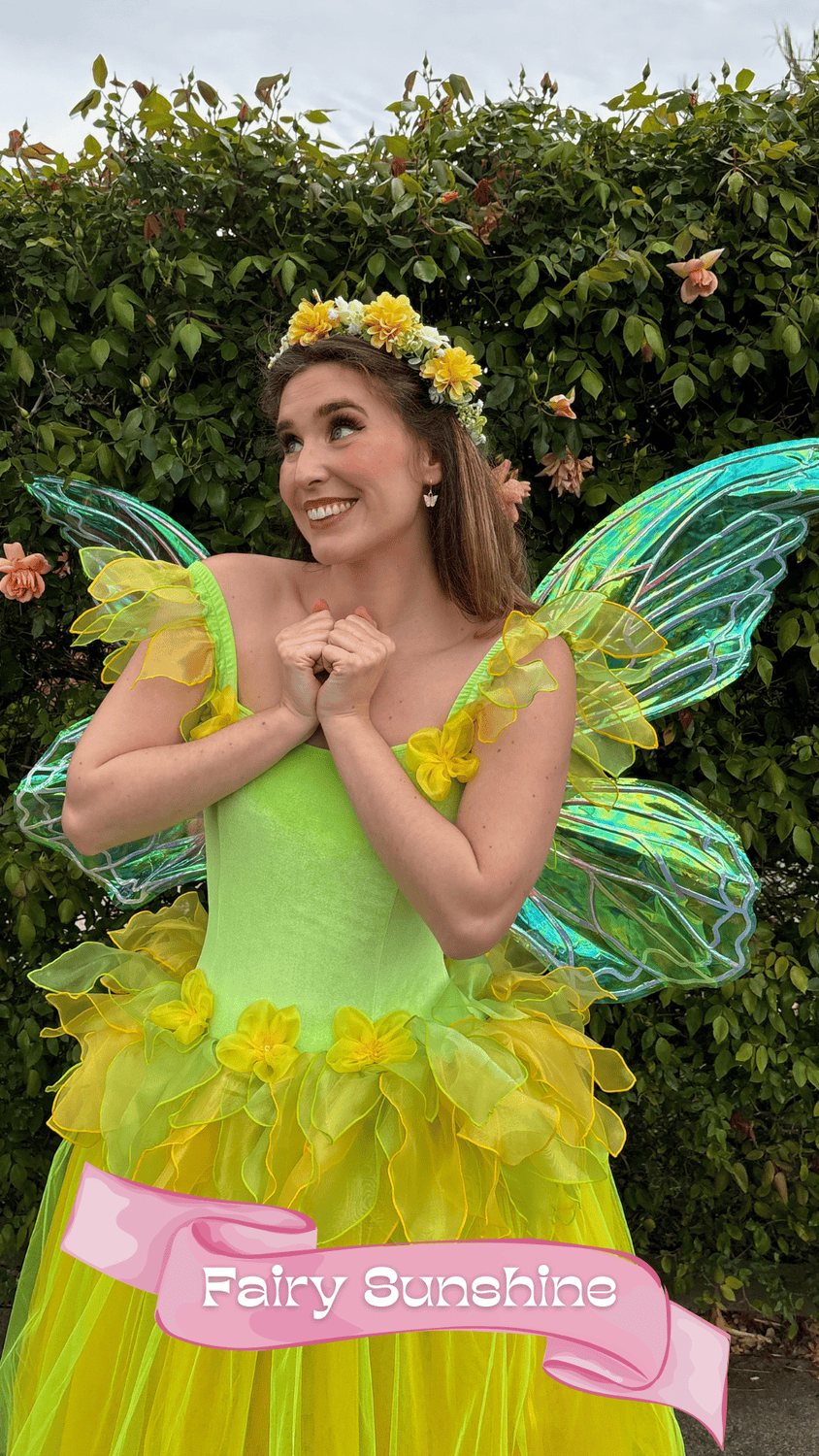 Woman dressed as a fairy fairy in a bright yellow and green costume with fairy wings, standing in front of a green leafy background and smiling.