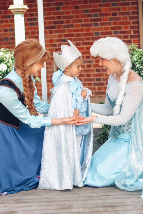 Three people in princess costumes smiling outdoors in front of a brick house, with greenery in the background.