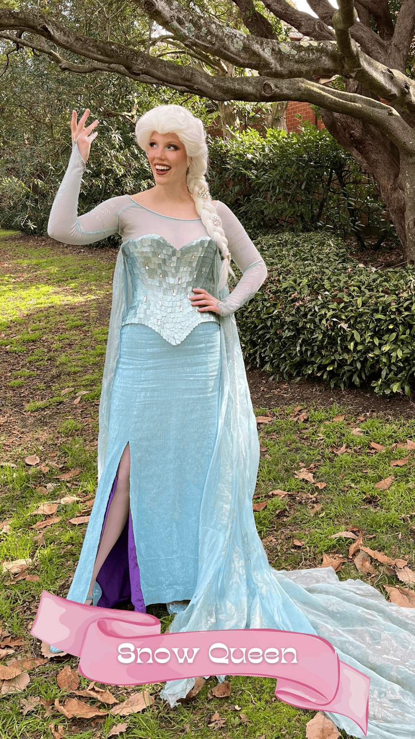 A woman dressed as the Snow Queen from a fairy tale, wearing a long, light blue gown with a high slit, standing outdoors under a tree with green leaves and bushes in the background. She has long, white hair styled in a braid and is smiling while hold