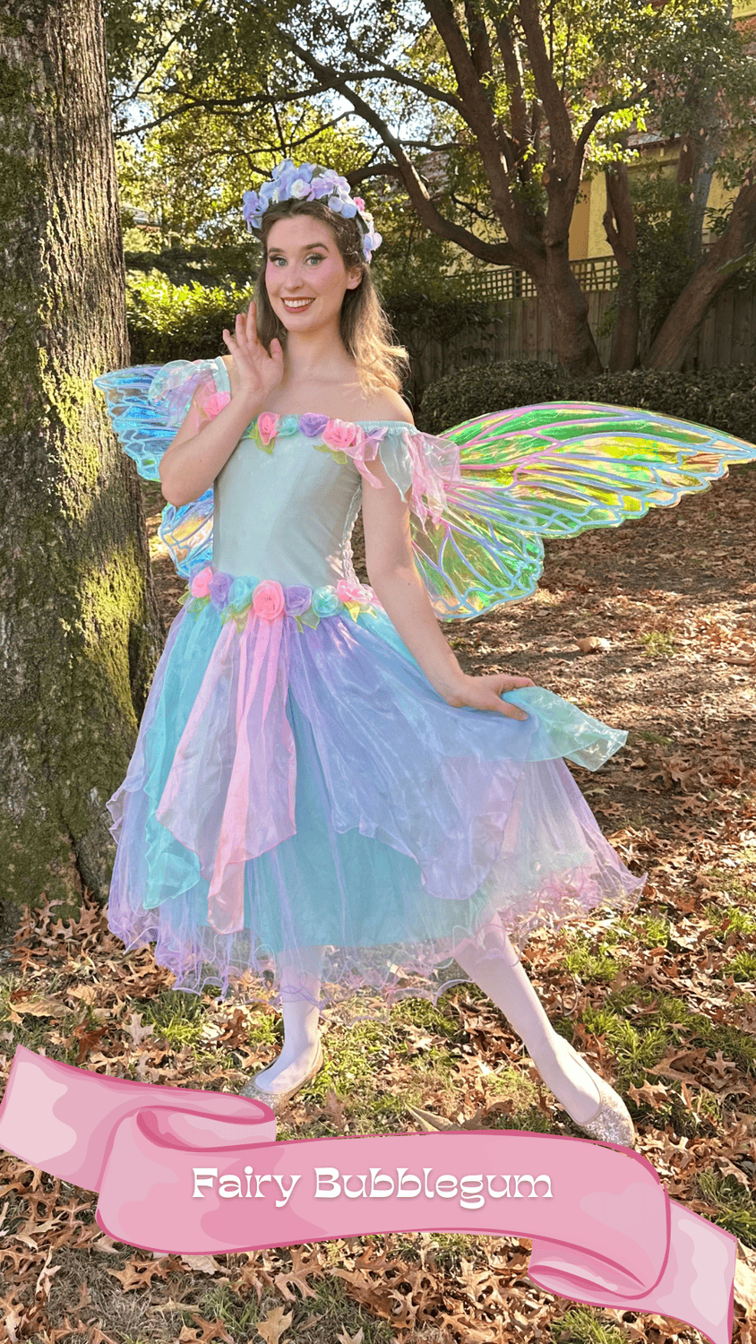 A woman dressed as a fairy with iridescent butterfly wings, wearing a pastel-colored dress with flower accents, a floral headpiece, standing outdoors in a wooded area during daytime.