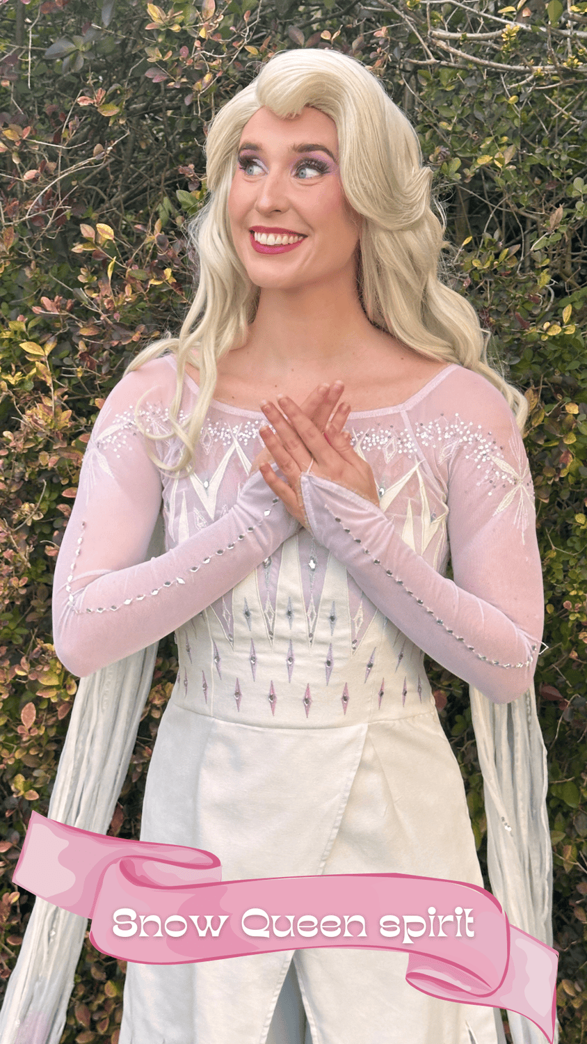 A woman dressed as a snow queen with long blond hair, silver and pink gown with intricate embroidery, standing in front of a bush with green and brown leaves, smiling and placing her hands on her chest. A pink banner with the text "Snow Queen spirit"