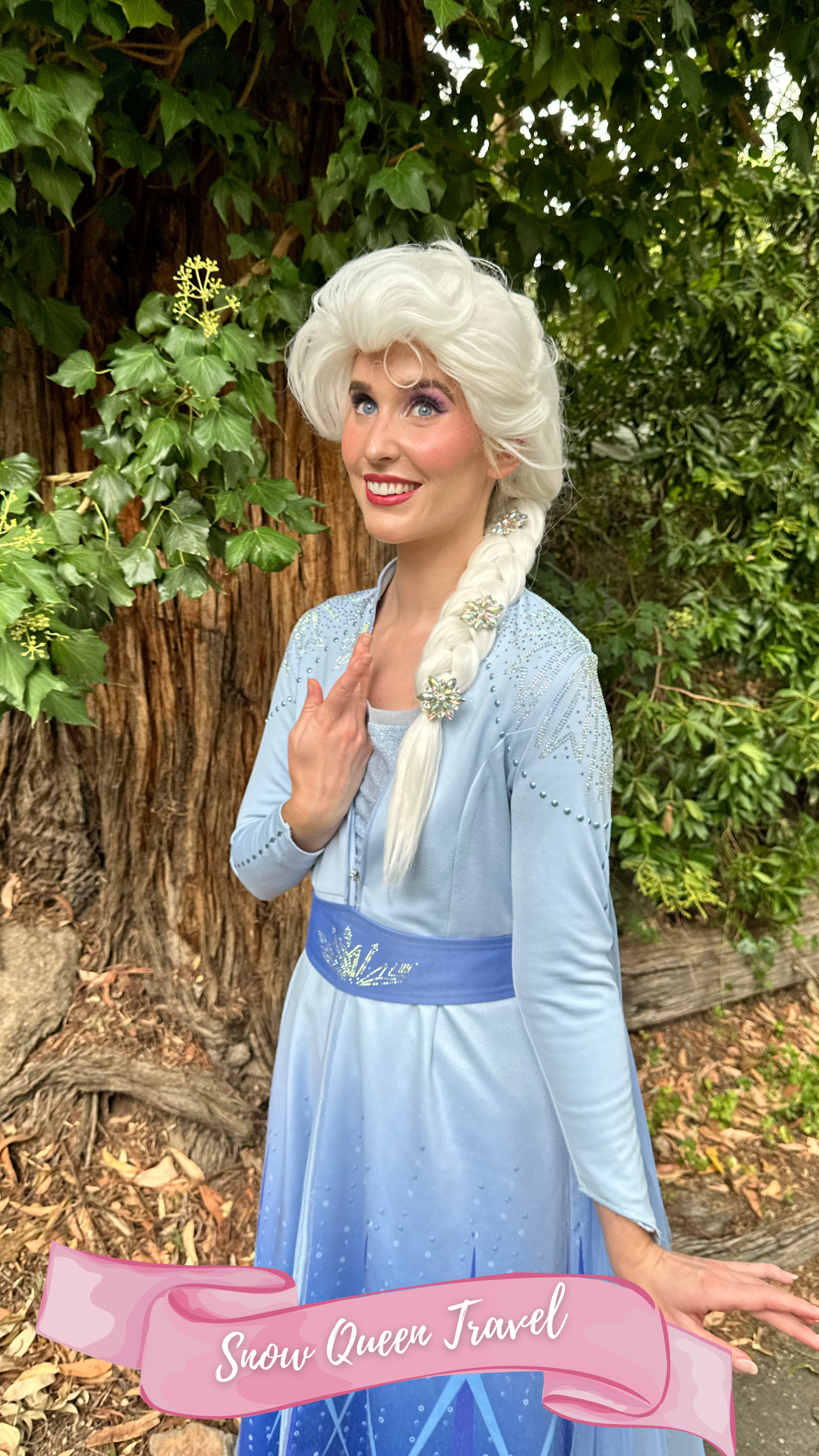 A woman dressed as the princess from the fairy tale Frozen, wearing a blue jacket, blue top, sparkly belt with snowflakes in her white hair, standing outdoors on a path with trees in the background.