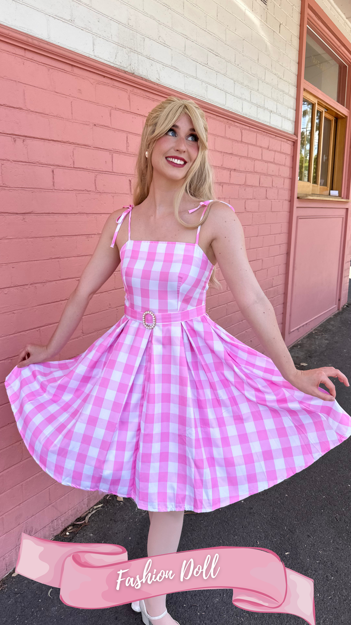 A woman standing in front of a pink building, wearing a pink and white checkered dress with shoulder ties, a matching pink belt with a pearl buckle, pink sunglasses on her head, and a pink hair scarf, smiling cheerfully.