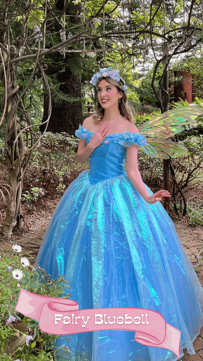 A woman dressed as a fairy princess in a blue dress with iridescent wings and a floral headband, standing outdoors in a garden surrounded by greenery and flowers.