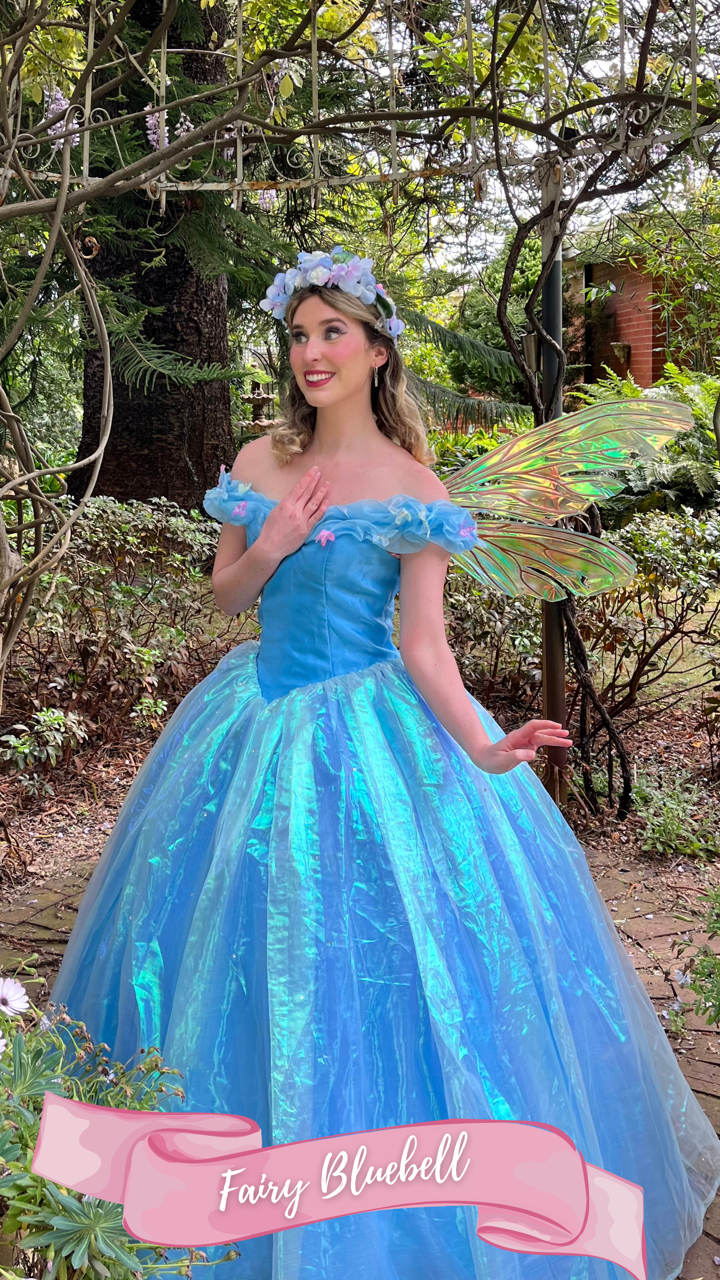 A woman dressed as a fairy princess in a blue dress with iridescent wings and a floral headband, standing outdoors in a garden surrounded by greenery and flowers.